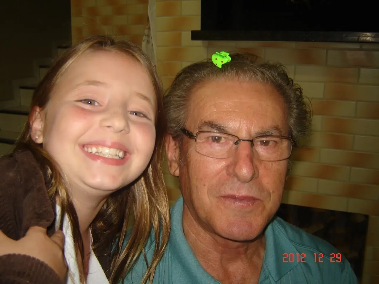 Uma jovem sorridente com cabelo castanho comprido e um homem mais velho de óculos e camisa azul, que tem um objeto verde no cabelo.