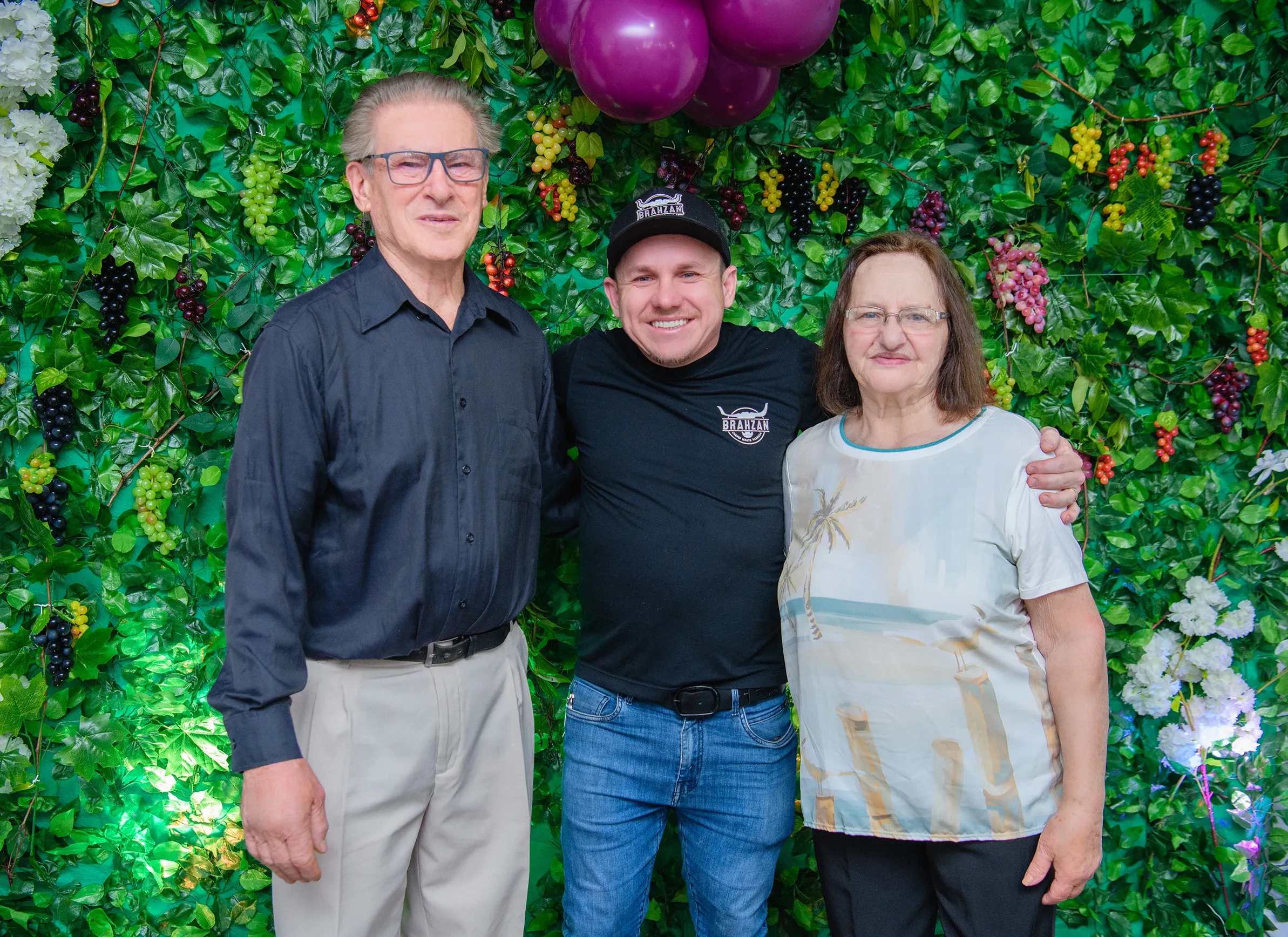 Três pessoas, dois idosos e um homem mais jovem no meio, sorriem para a câmera em frente a uma parede coberta por folhagens verdes e cachos de uva roxos, verdes e vermelhos; um buquê de balões roxos paira no canto superior esquerdo e flores brancas e luzes cintilantes adornam a parte inferior.