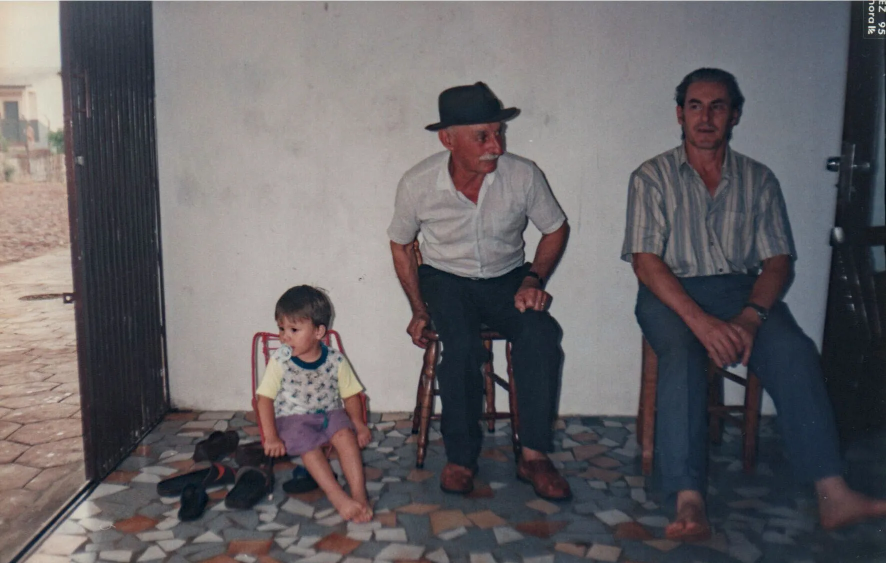 Três homens sentados, um menino descalço e dois homens mais velhos sentados em bancos, dentro de casa com a porta aberta para o exterior.