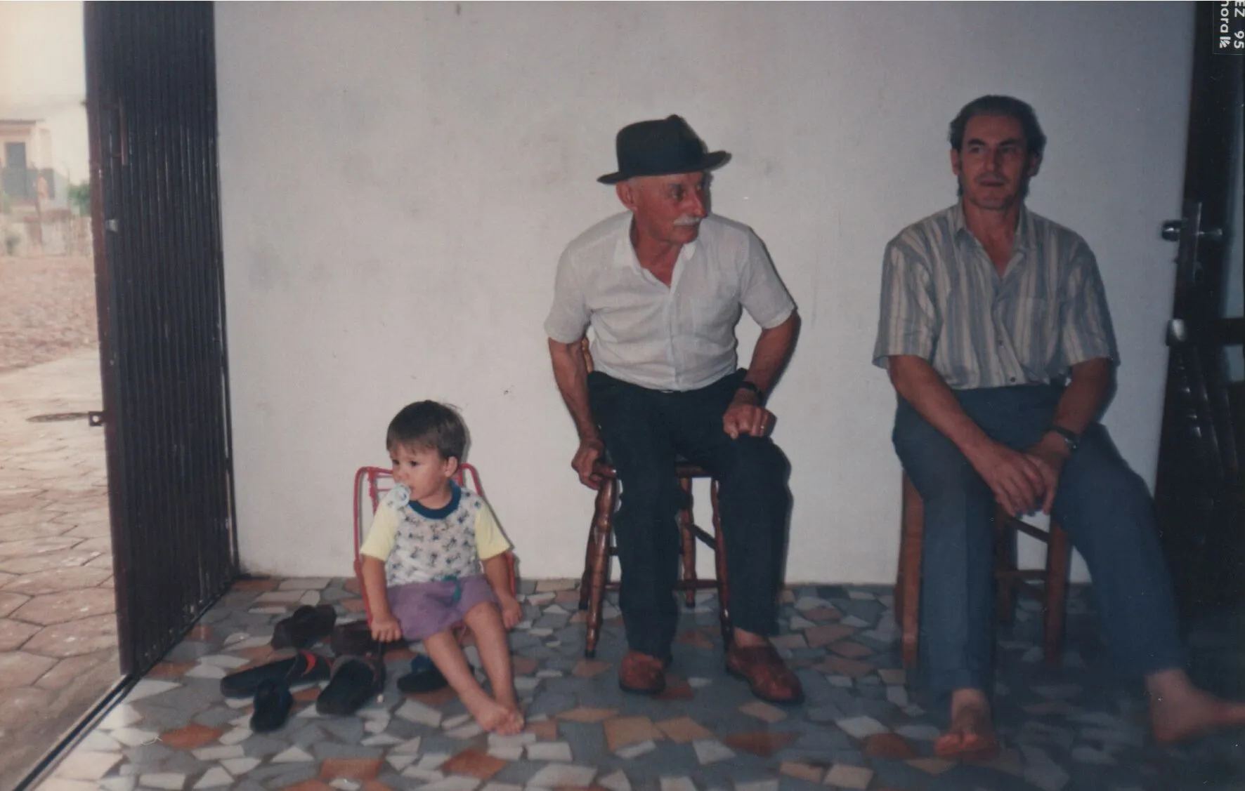 Três homens sentados, um menino descalço e dois homens mais velhos sentados em bancos, dentro de casa com a porta aberta para o exterior. (Original)