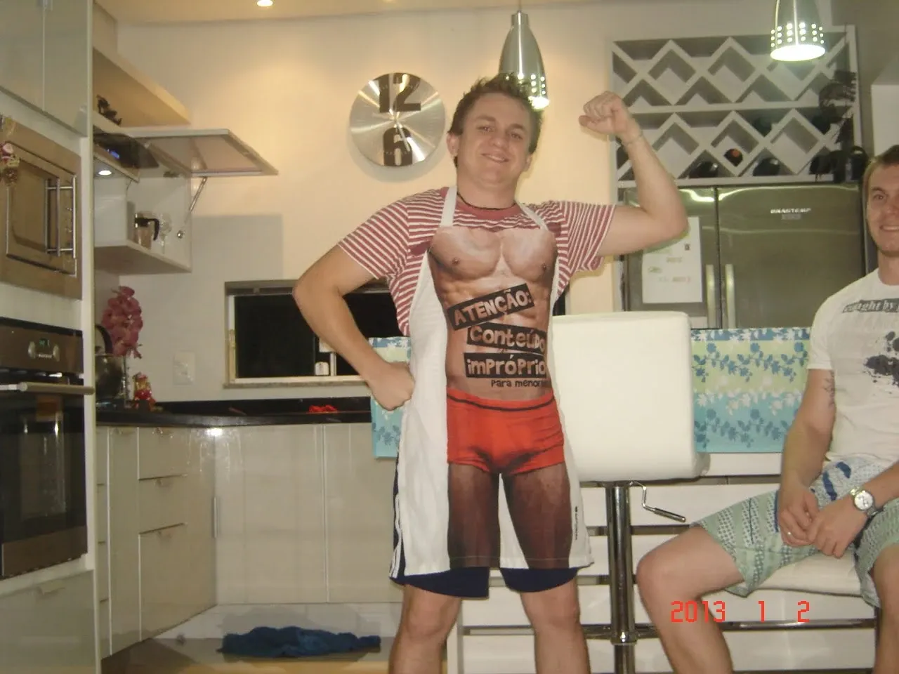 Um homem de camiseta de listras e avental com um desenho de torso musculoso posa com os braços flexionados na cozinha enquanto outro homem se senta à direita.