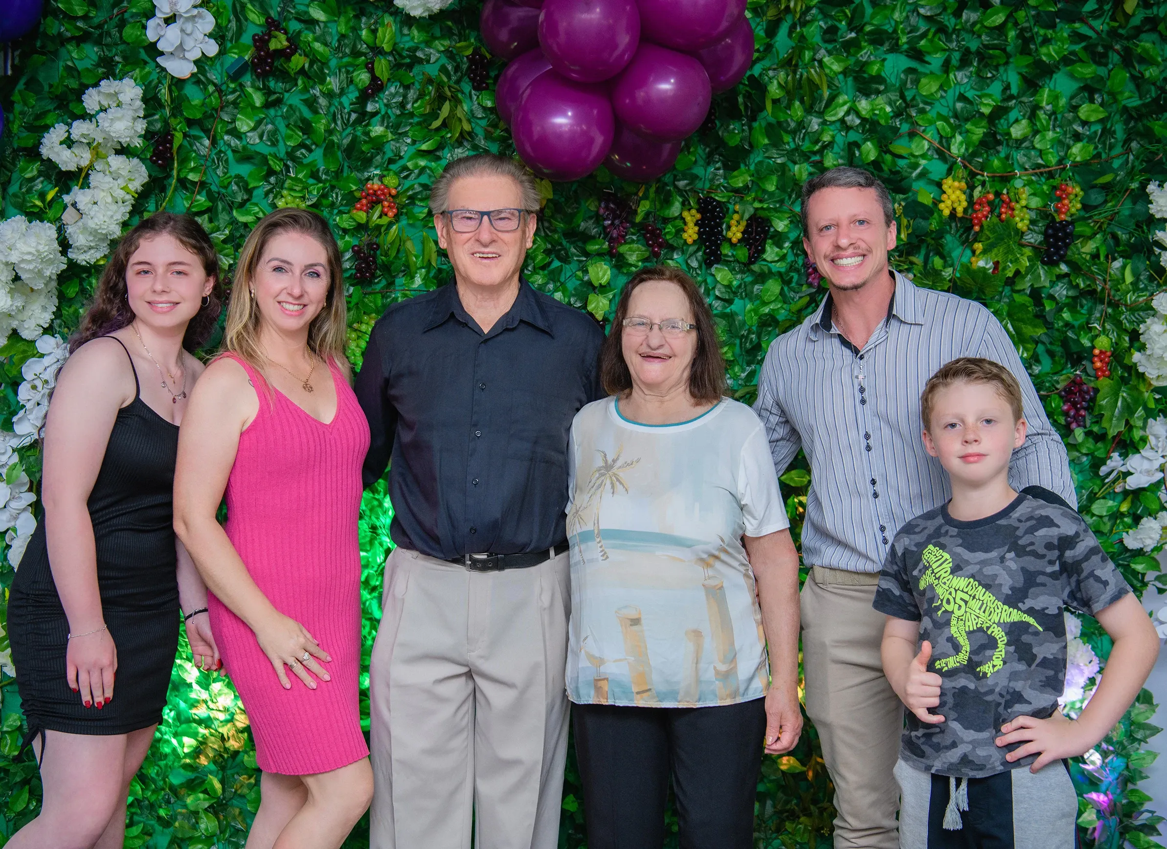 Uma família sorridente de seis pessoas posa para uma foto em frente a uma parede de vegetação exuberante, decorada com flores brancas e roxos.