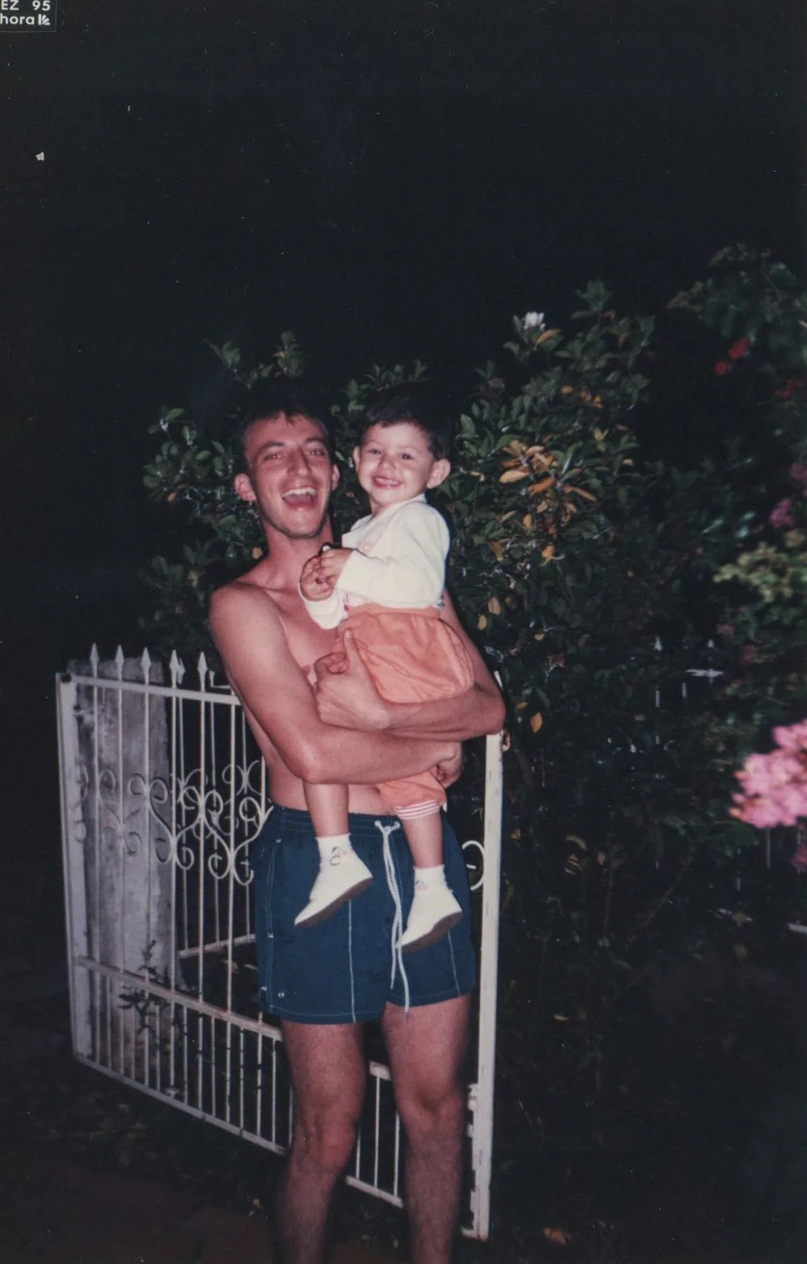 Homem sem camisa em shorts azuis segura uma criança sorridente em roupas pêssego e creme na frente de um portão branco e folhagem escura à noite.