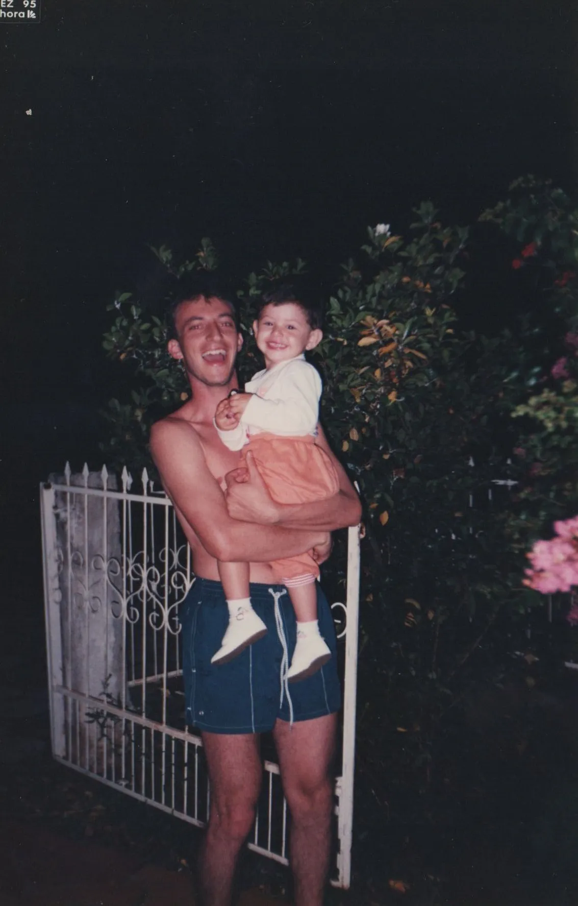 Homem sem camisa em shorts azuis segura uma criança sorridente em roupas pêssego e creme na frente de um portão branco e folhagem escura à noite. (Original)