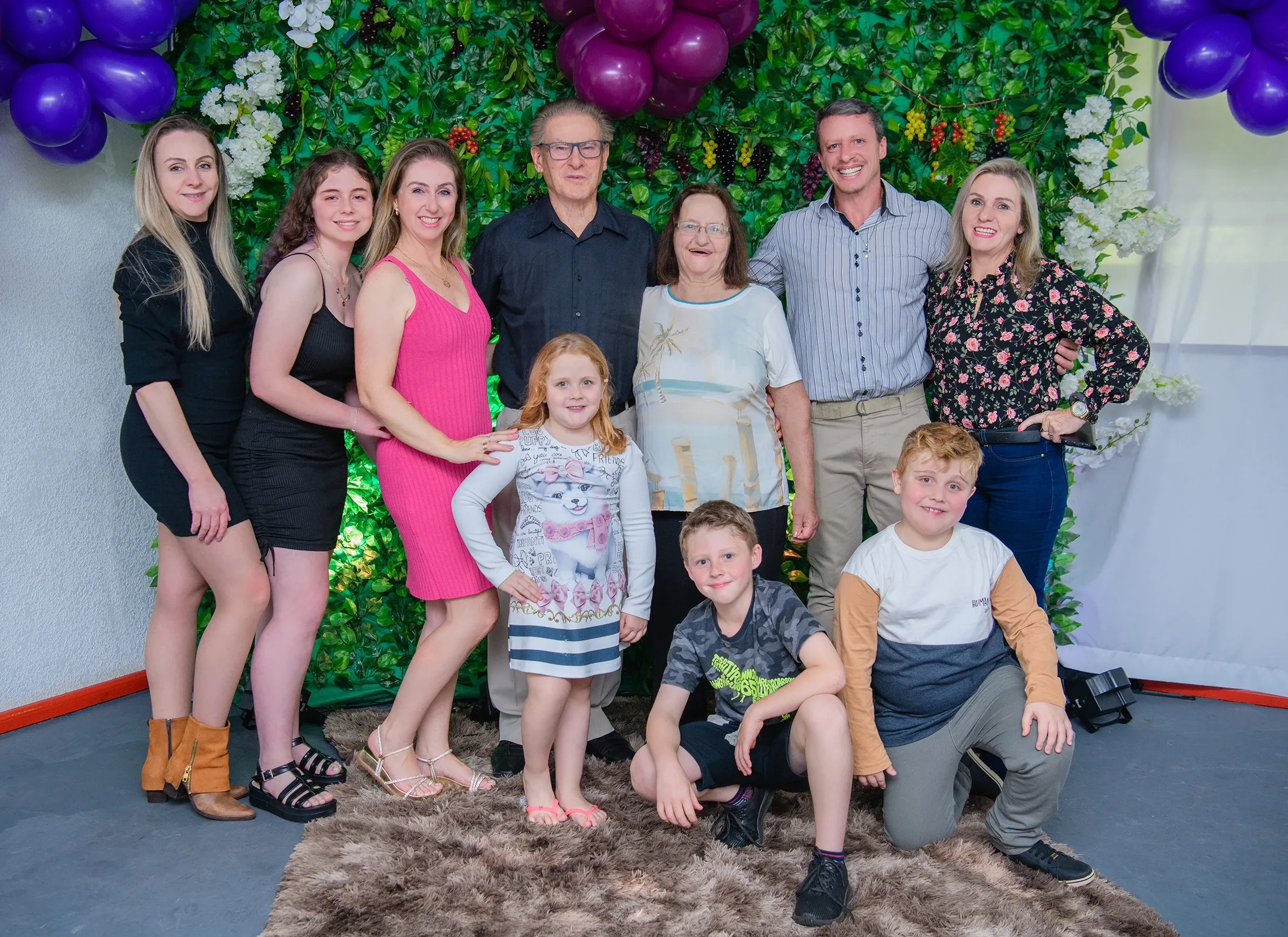 Um grupo de dez pessoas, incluindo avós, pais e crianças de diferentes idades, posa junto em frente a uma parede decorada com folhagem verde e flores brancas, balões roxos visíveis no canto superior esquerdo e balões pretos no canto superior direito.