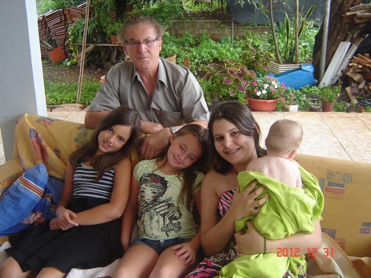 Um homem idoso sentado em um pátio externo atrás de quatro meninas sentadas em um sofá, com a garota da direita segurando um bebê.