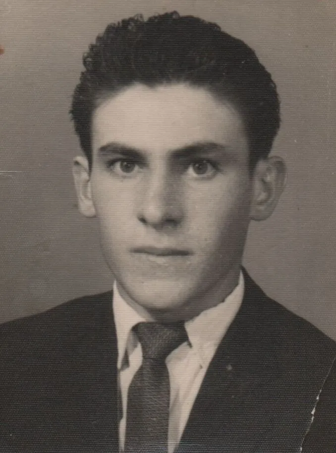 Um jovem sério com cabelo escuro e olhos castanhos, vestindo um terno cinza escuro com uma camisa branca e gravata azul-marinho, olhando diretamente para frente com uma expressão neutra. (Original)