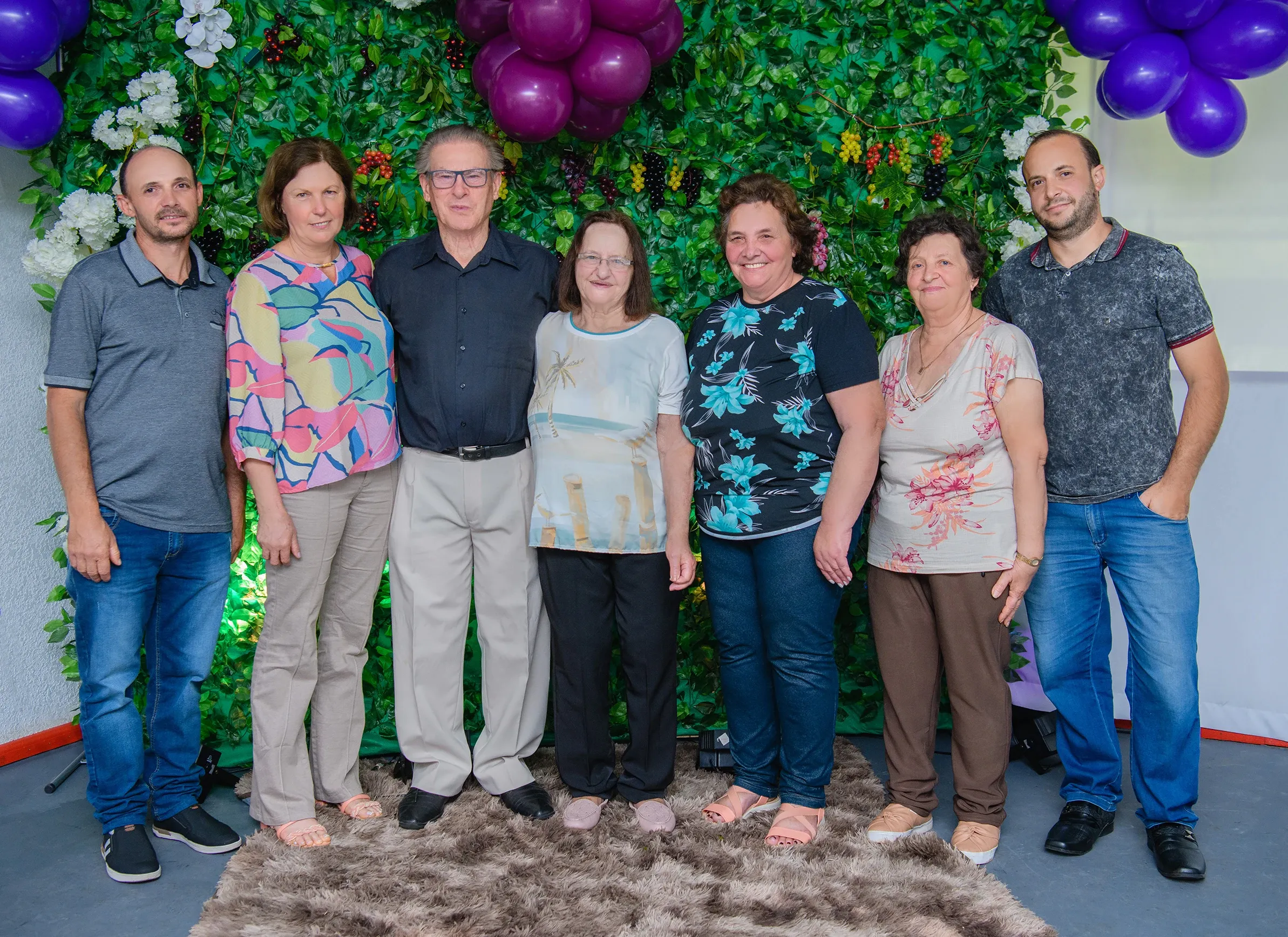 Sete adultos, três homens e quatro mulheres, posam para uma foto em pé na frente de um fundo floral e folhoso com balões roxos acima.