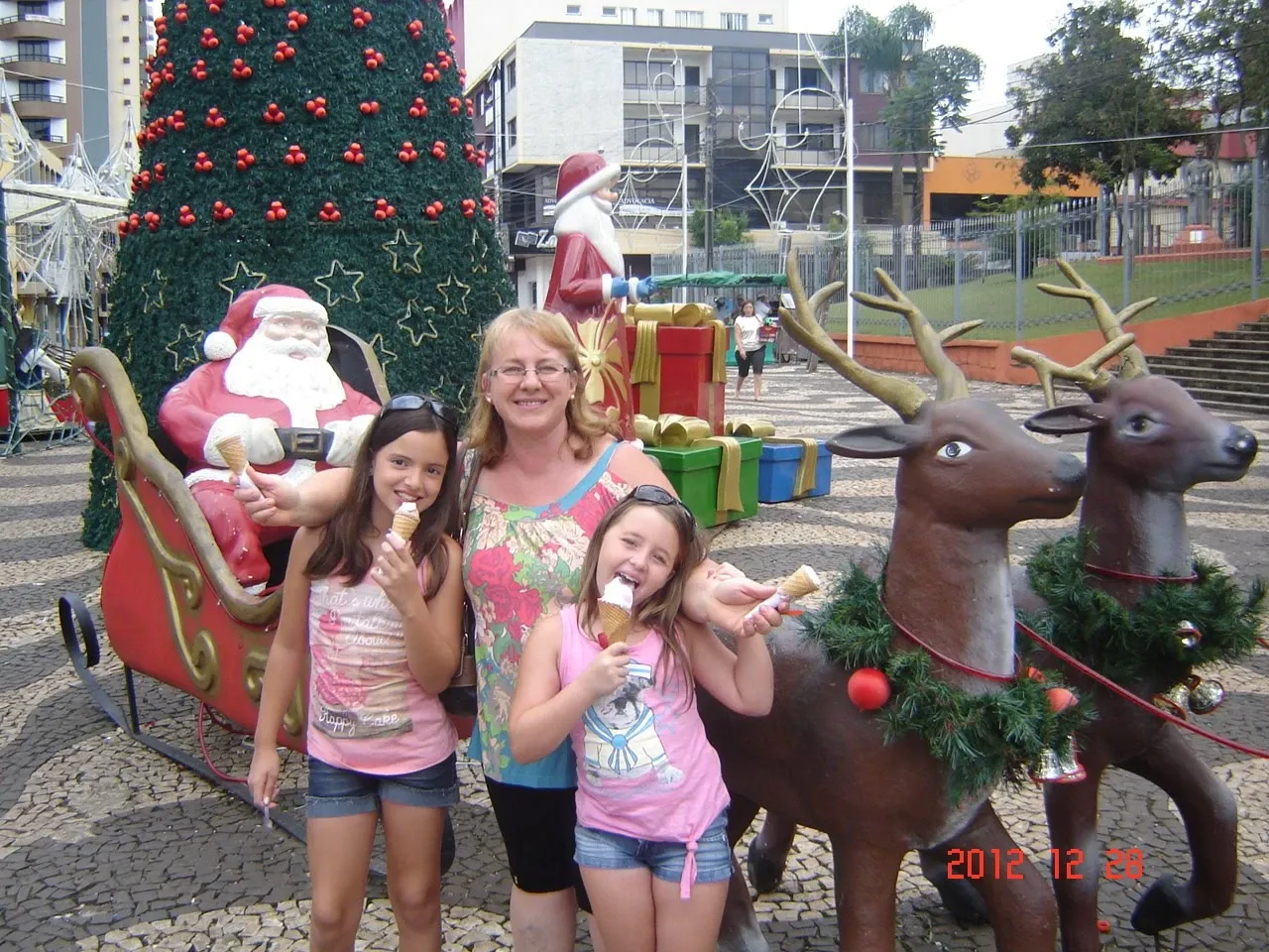 Três mulheres e um Papai Noel de tamanho real em um trenó comendo sorvete ao lado de duas estátuas de rena.