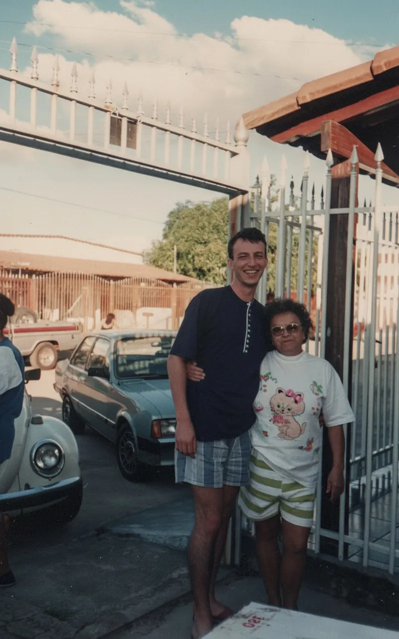 Um homem e uma mulher idosa abraçados e sorrindo, de pé na frente de um portão de metal, com carros e casas em segundo plano.