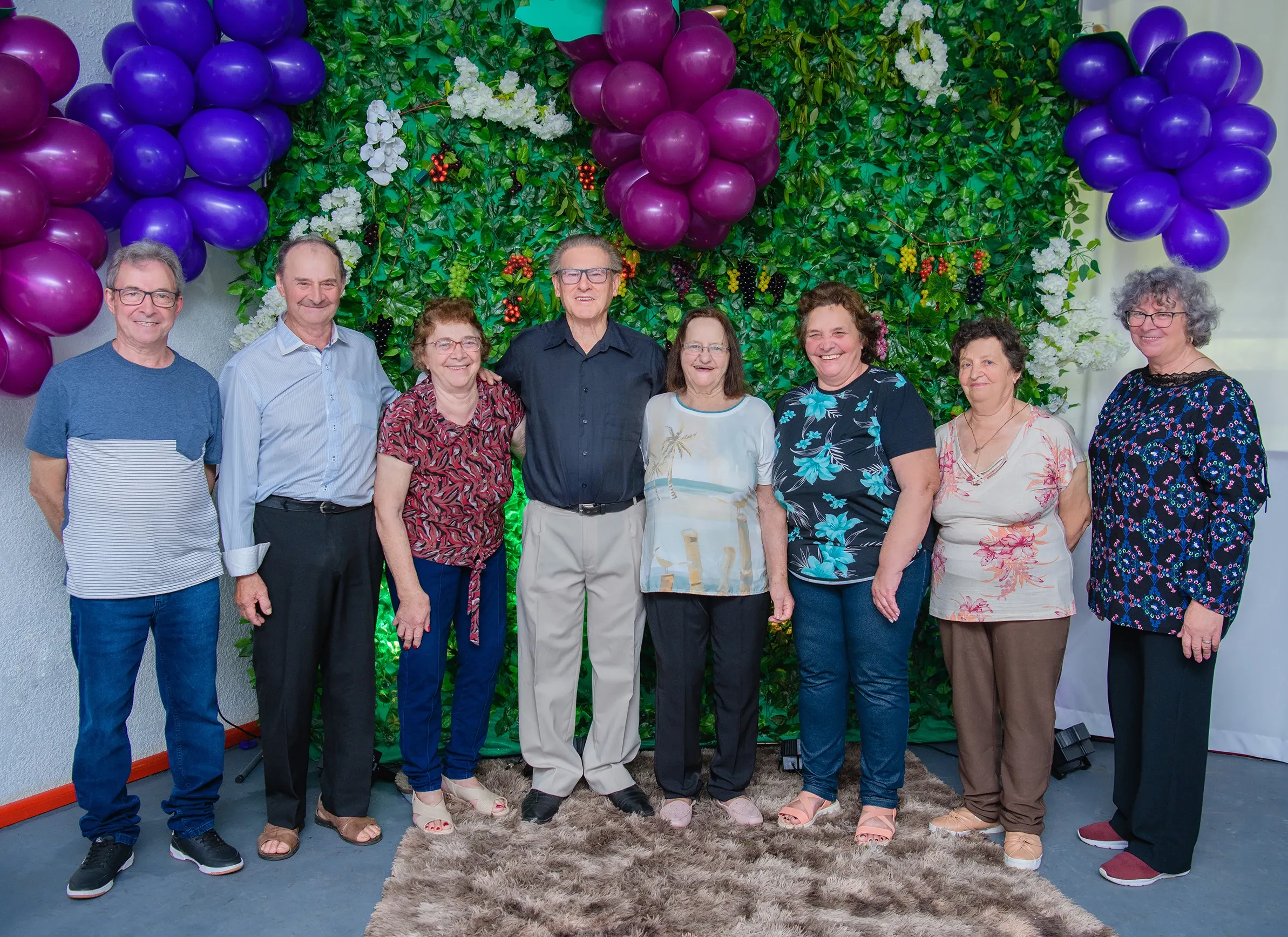 Um grupo de pessoas mais velhas, quatro homens e quatro mulheres, está em frente a uma parede verde decorada com balões roxos, um tapete felpudo marrom debaixo deles.