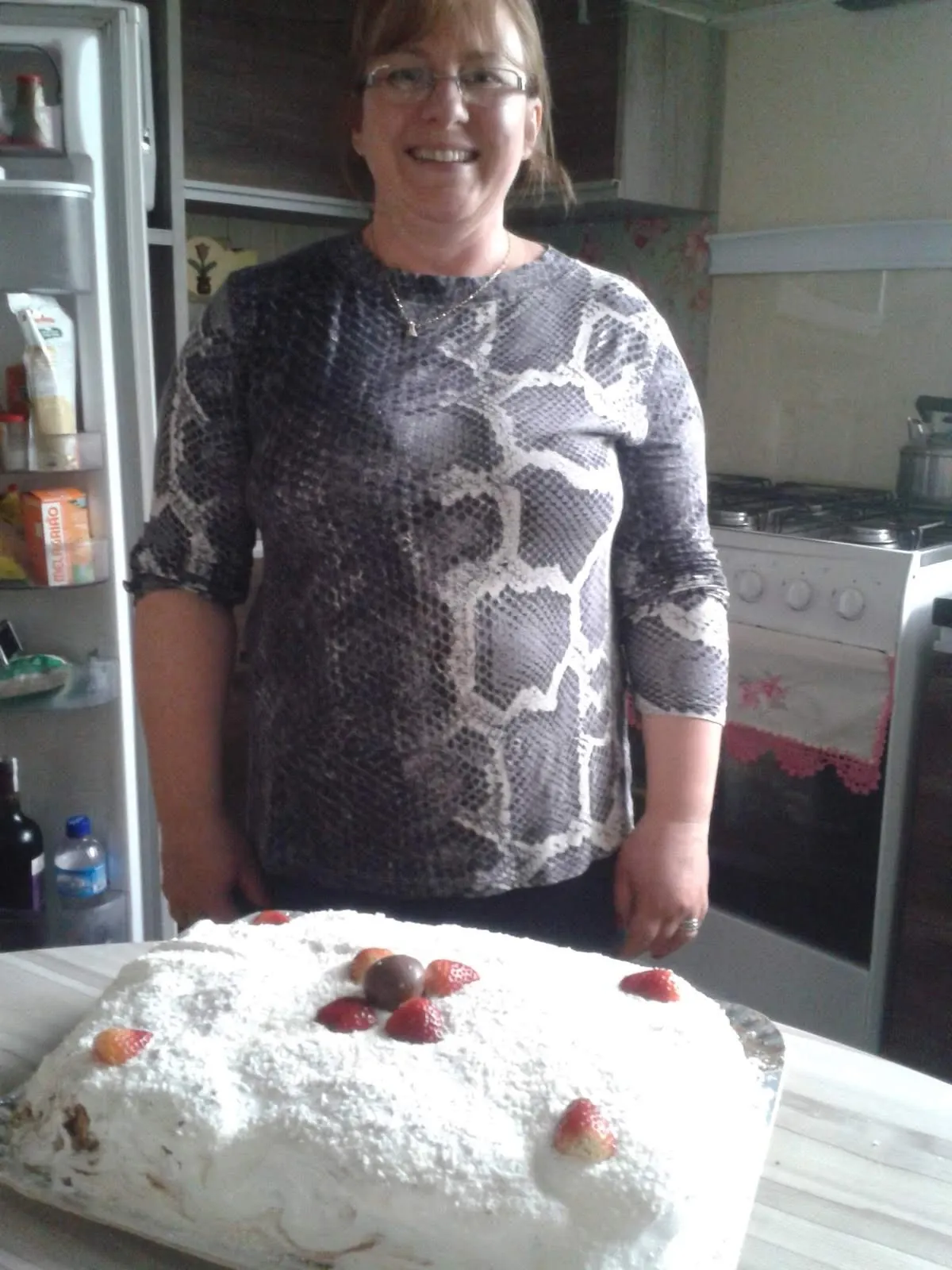 Uma mulher de óculos sorri para a câmera atrás de um bolo coberto de branco com morangos e uma pequena bola de chocolate, em uma cozinha.