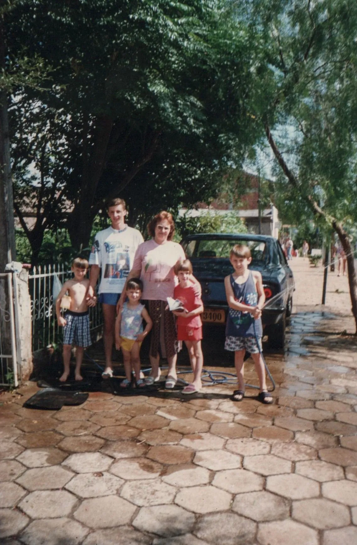 Um grupo de cinco pessoas – uma mulher, um homem e três crianças – está em frente a uma casa com árvores e um carro estacionado na entrada.