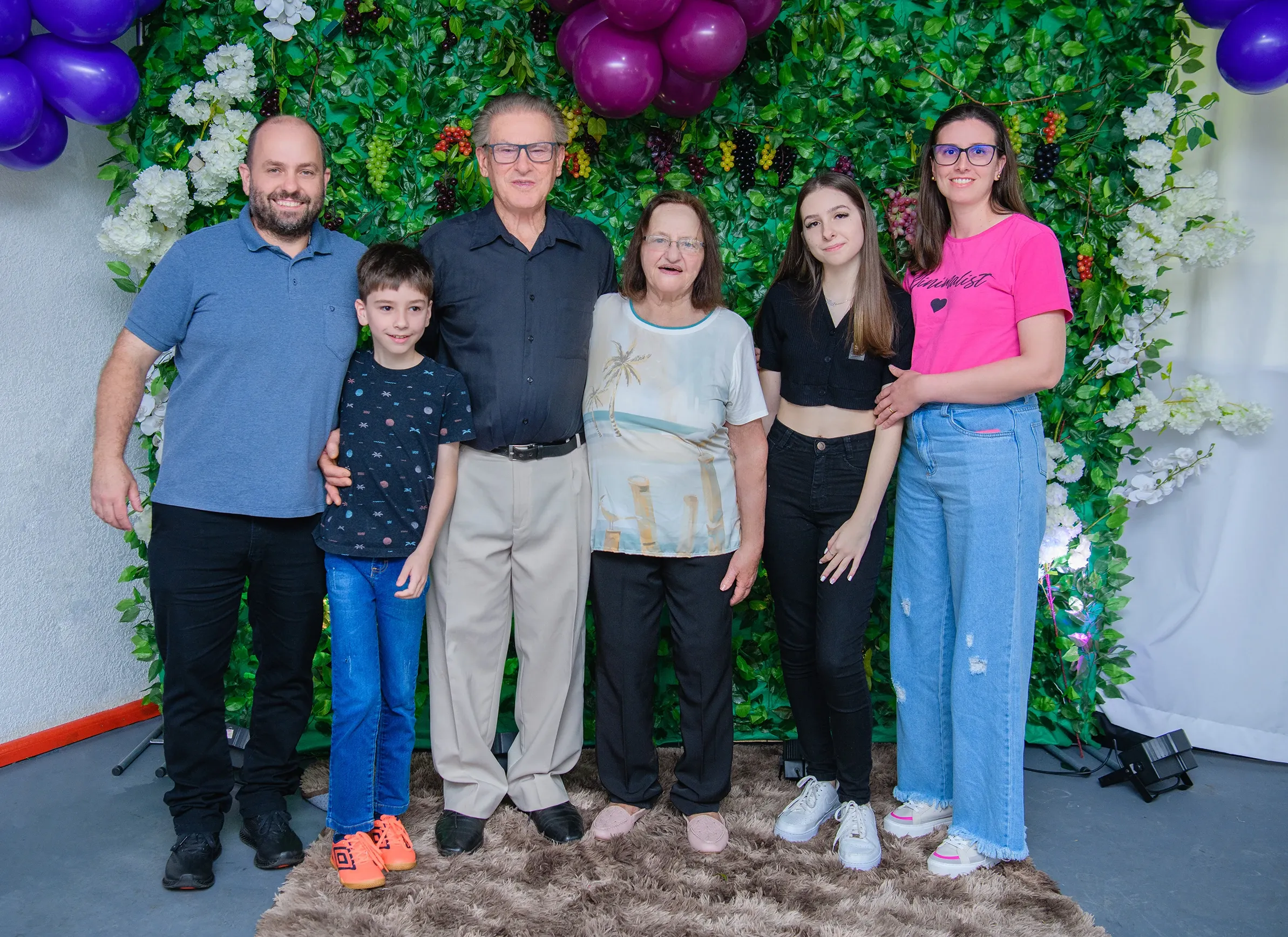 Seis pessoas de diferentes idades em frente a uma parede de folhagem verde e flores brancas, com balões roxos no canto superior esquerdo e superior central.