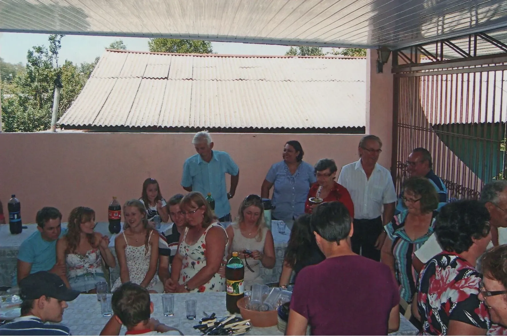Grupo de adultos e adolescentes reunidos sob um pátio coberto, com pessoas sentadas a uma mesa à esquerda e outras de pé ao fundo e à direita, celebrando uma reunião familiar num ambiente exterior casual.