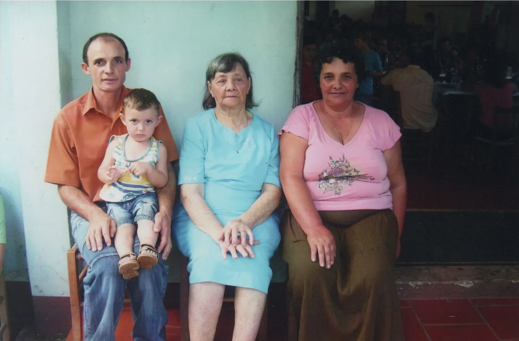 Quatro pessoas sentadas lado a lado em um ambiente interno. Um homem de camisa laranja segura uma criança no colo. À esquerda, uma mulher de vestido azul claro senta-se com as mãos cruzadas, e à direita, outra mulher de camiseta rosa e saia marrom senta-se com as mãos nos joelhos.