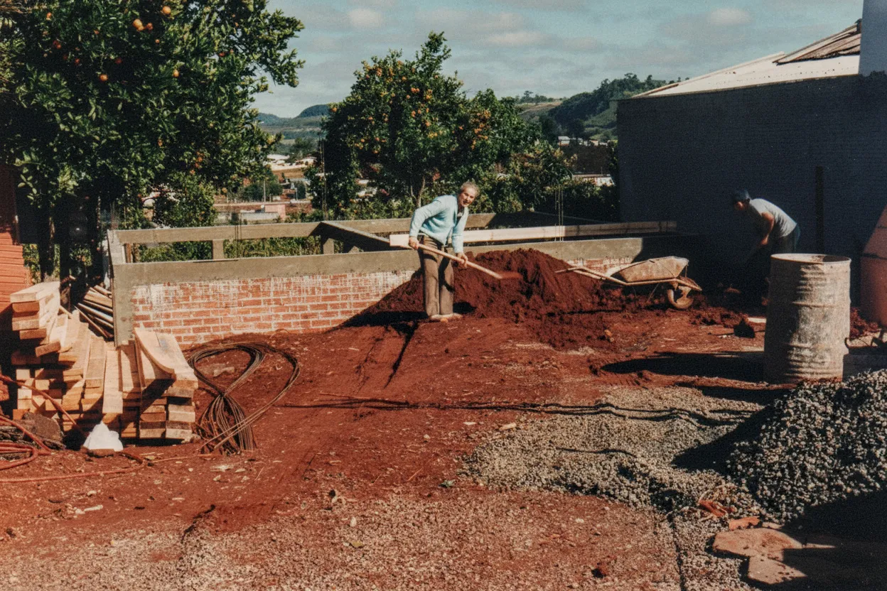 Dois homens trabalham em uma obra de construção com tijolos e terra vermelha, sob um céu parcialmente nublado, com árvores e casas ao fundo.