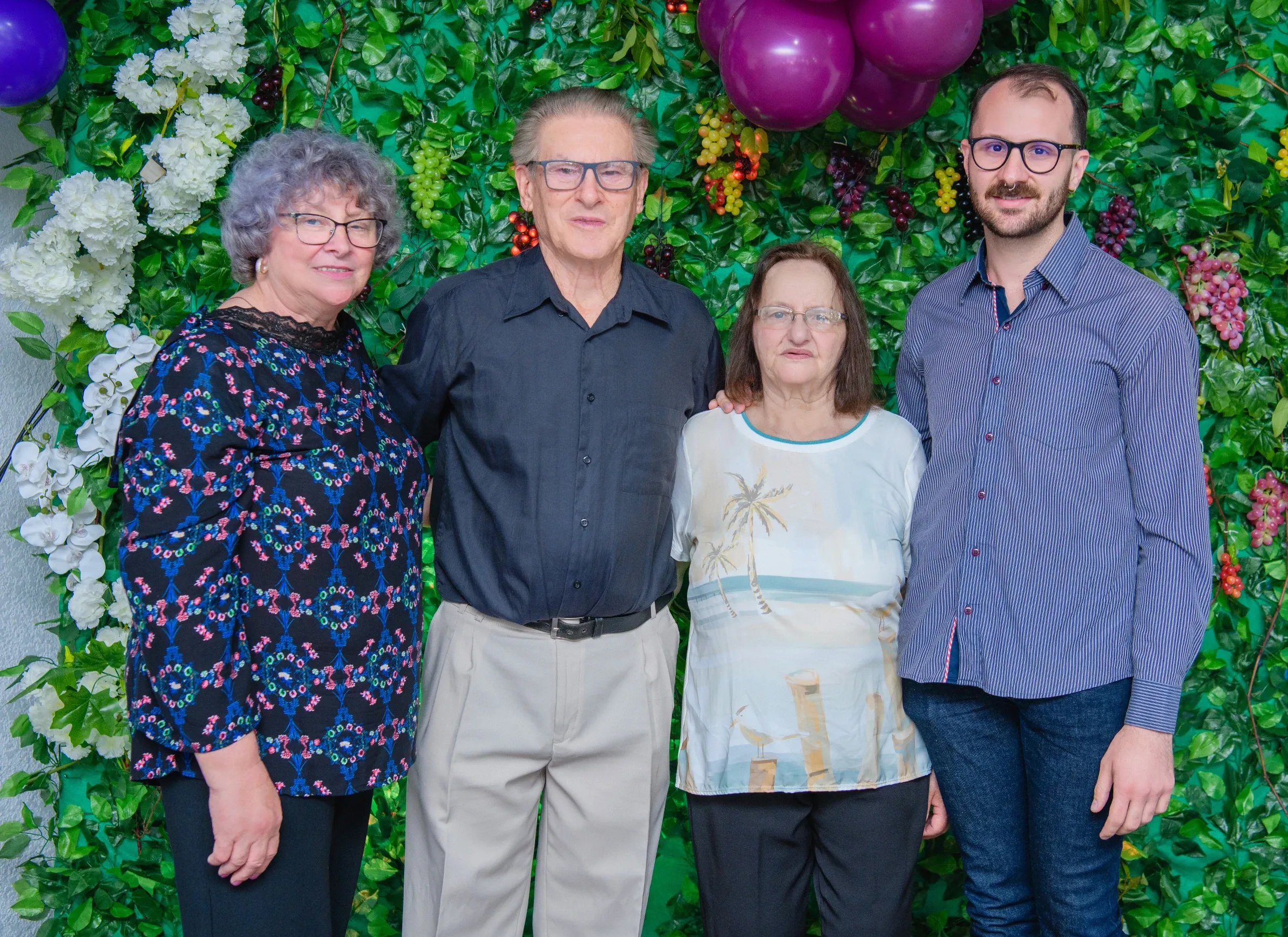 Quatro pessoas, duas mulheres e dois homens de diferentes idades, estão em pé e posando para a câmera em frente a uma parede coberta por plantas verdes e flores brancas e roxas, com balões roxos no canto superior direito e esquerdo.