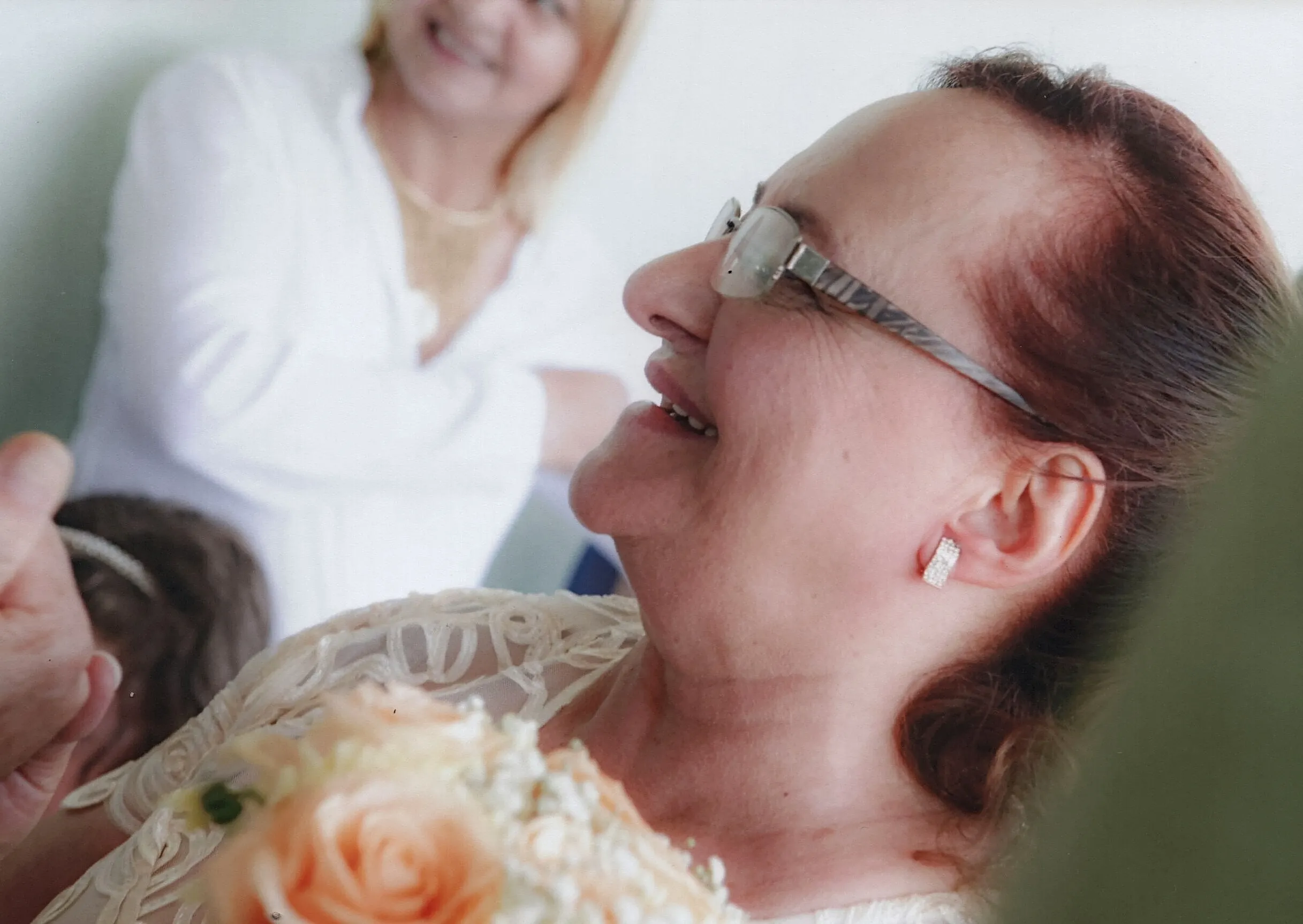 Uma mulher sorridente com óculos e cabelo ruivo está em primeiro plano, com um buquê de rosas cor de pêssego em suas mãos, enquanto outra mulher sorridente com cabelo loiro está desfocada ao fundo.