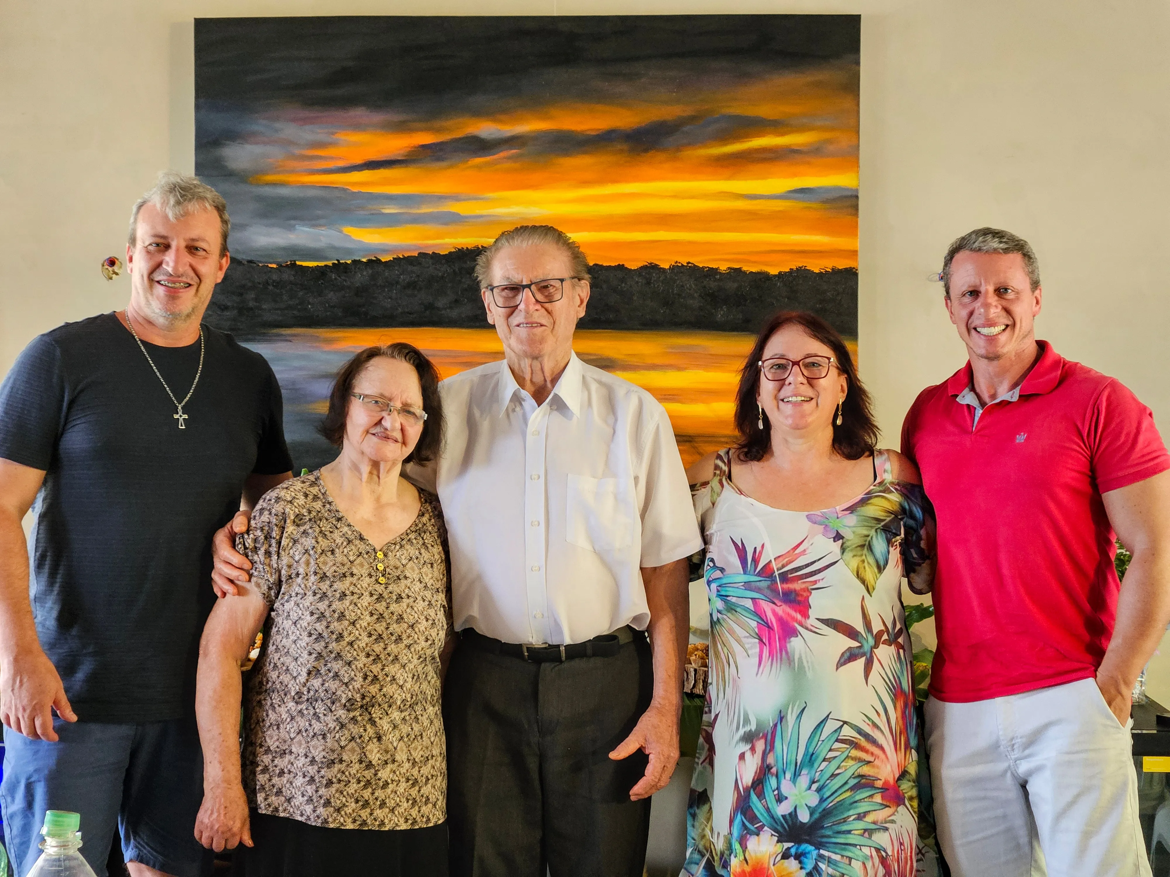 Cinco pessoas, possivelmente uma família, posam para uma foto em pé, sorrindo em frente a uma pintura abstrata.