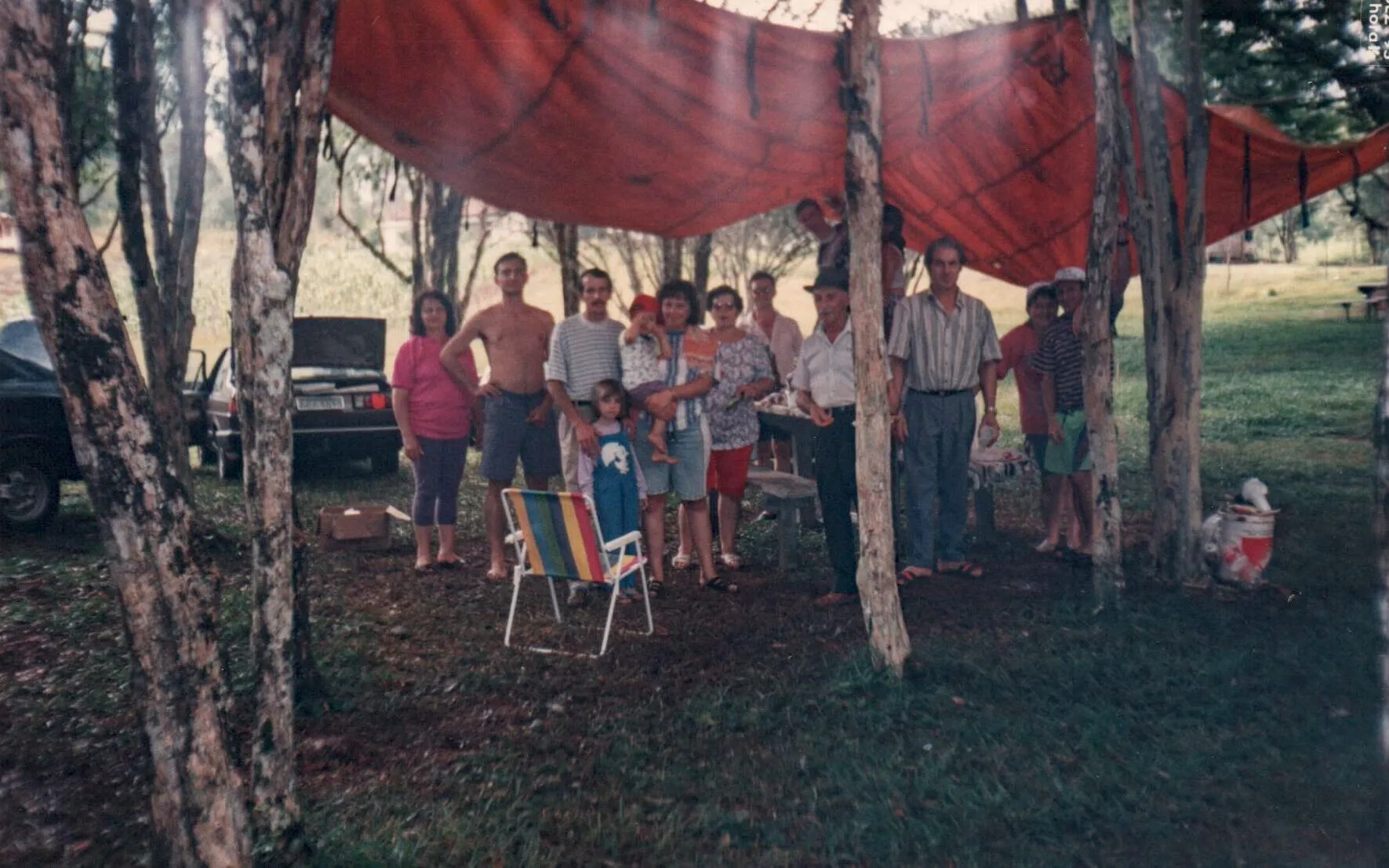 Um grupo de pessoas sob uma lona vermelha esticada entre árvores, com dois carros estacionados na grama atrás deles.