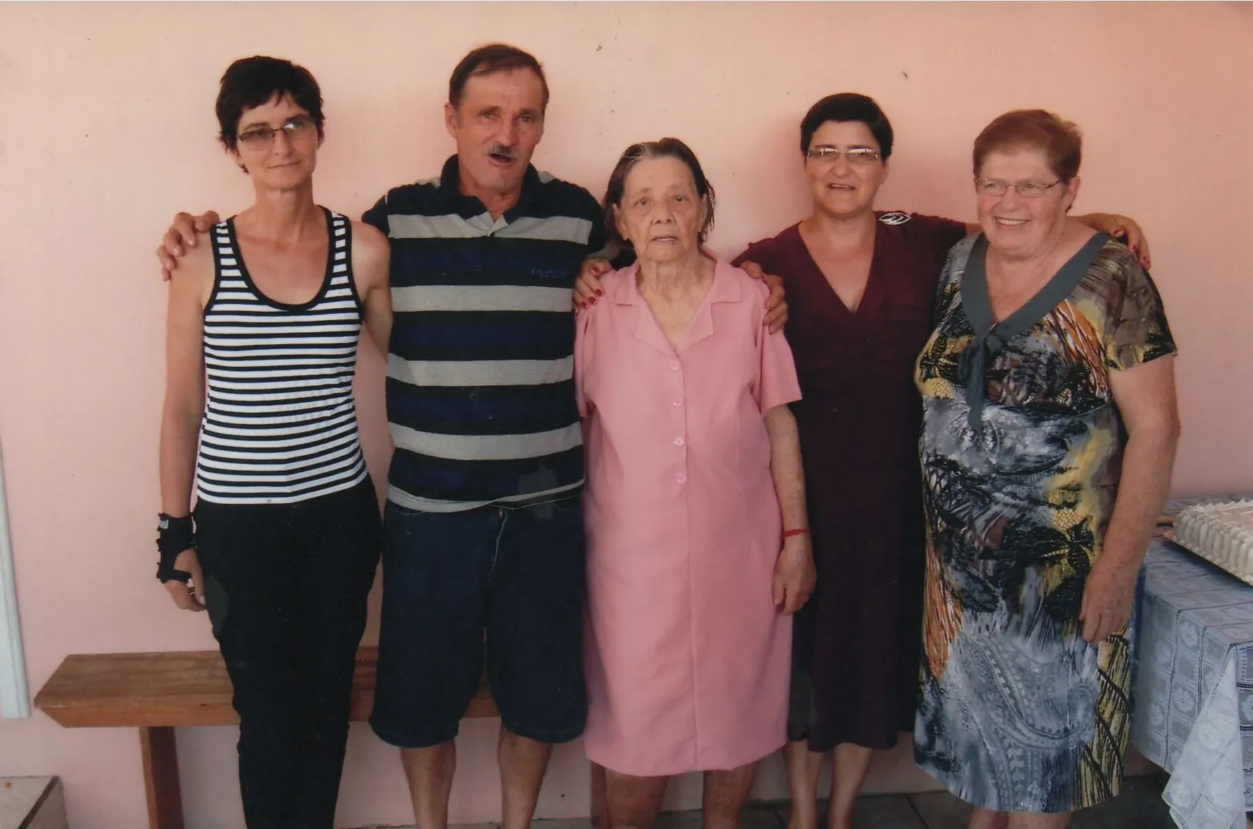 Cinco pessoas, possivelmente uma família de três irmãs, um homem e uma senhora idosa, em pé lado a lado na frente de uma parede rosa.