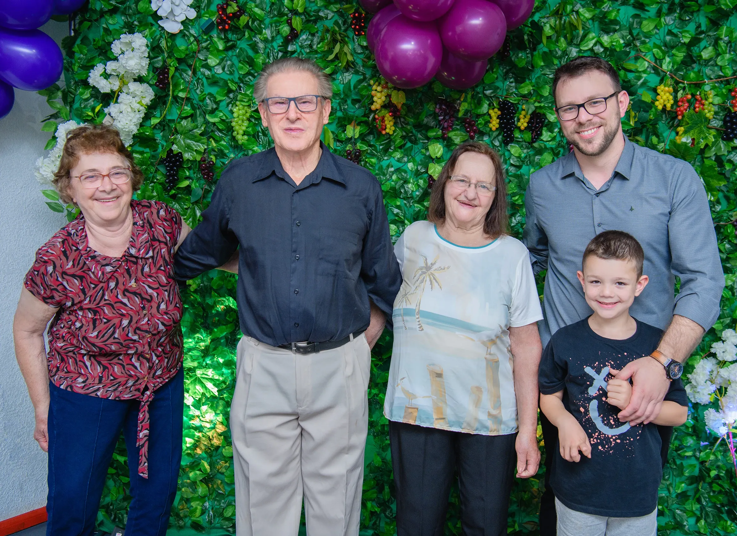 Cinco pessoas sorriem para a câmera em frente a uma parede verde decorada com folhagens e flores.