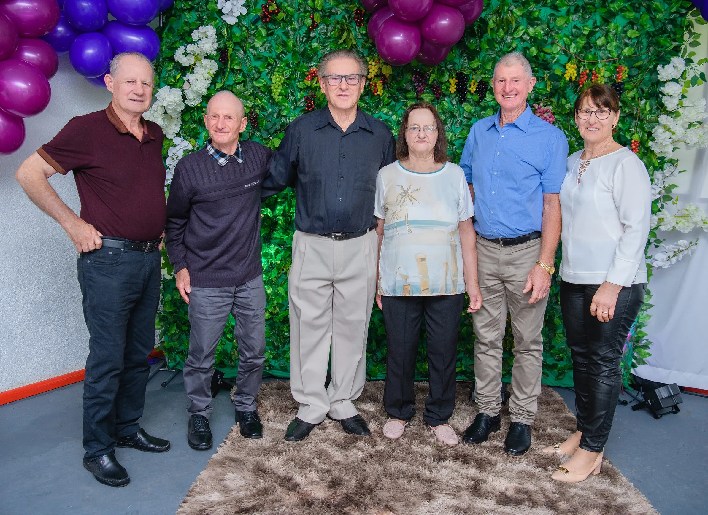 Seis pessoas adultas, três homens de cada lado de um casal e uma mulher, posam contra uma parede verde com folhagem, flores brancas e balões roxos.
