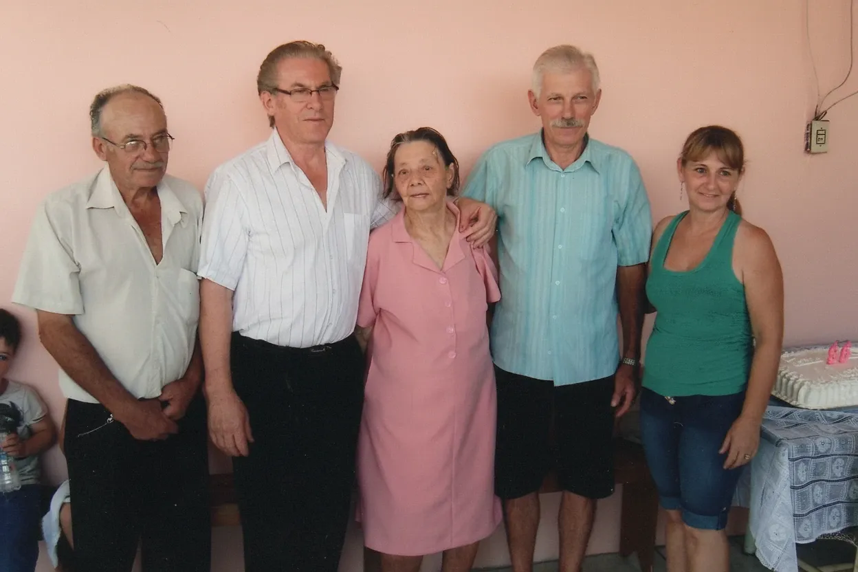 Cinco adultos posam para uma foto, em pé da esquerda para a direita: um homem idoso de camisa branca, um homem de óculos e camisa branca listrada, uma mulher idosa de vestido rosa, um homem de bigode e camisa azul, e uma mulher sorridente de regata verde e jeans. Um bolo de aniversário rosa está sobre uma mesa à direita.