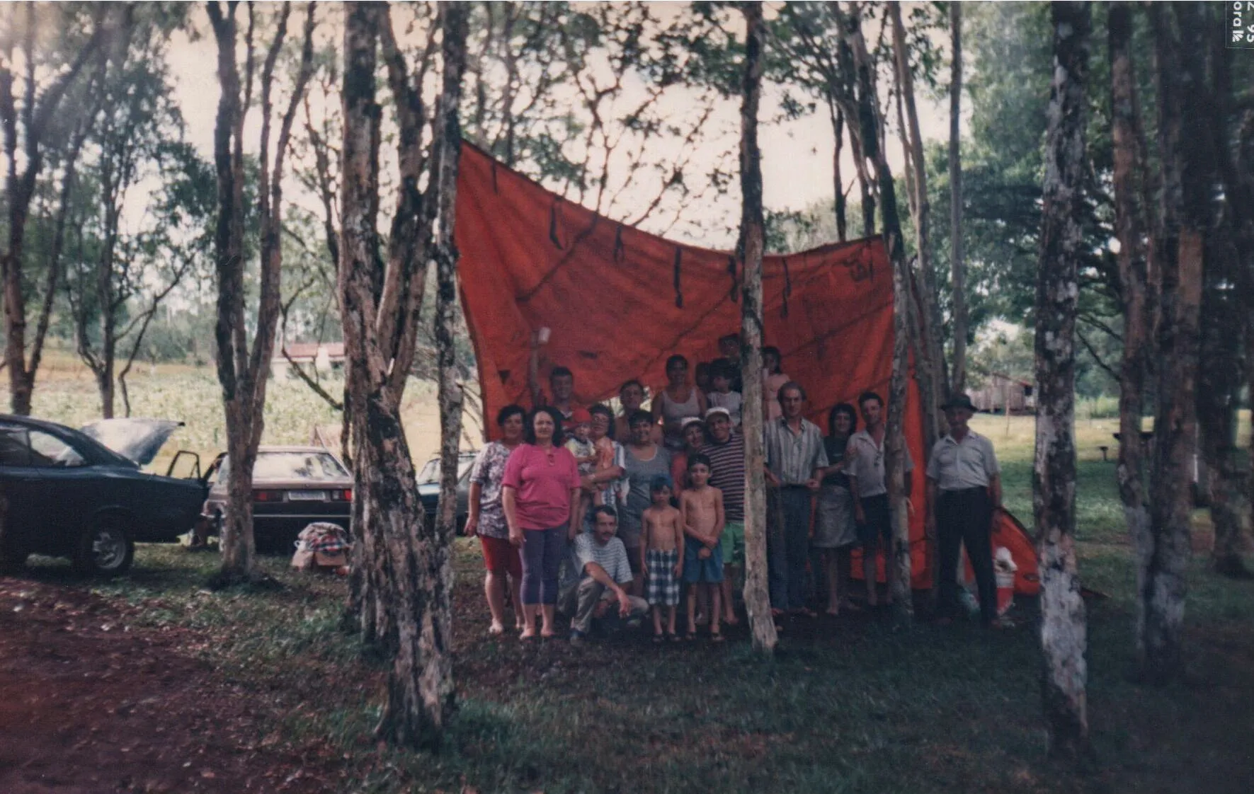 Um grande grupo de pessoas sorridentes, de todas as idades, posa ao ar livre sob um dossel vermelho improvisado feito de um pano grande, cercado por árvores, e carros antigos podem ser vistos ao fundo.