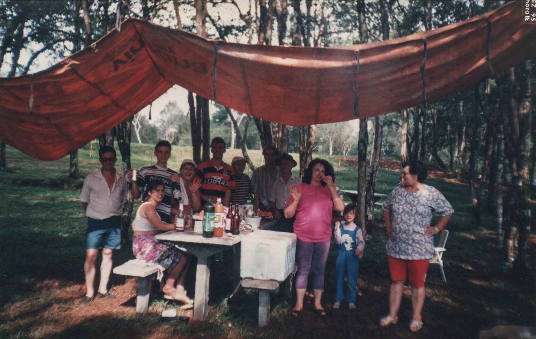 Um grupo de dez pessoas de diferentes idades, com dois homens e uma mulher sentados em um banco e o restante em pé, posam para uma foto em um acampamento sob uma lona laranja que serve de tenda.
