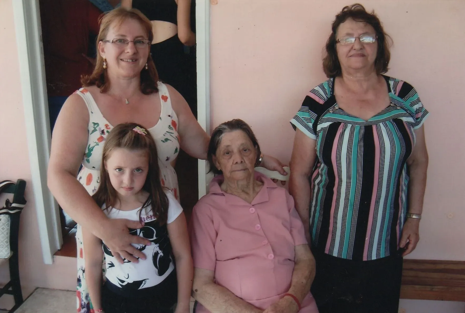 Cinco mulheres em diferentes gerações posam juntas; uma mulher mais velha sentada em uma cadeira rosa no centro é cercada por duas mulheres em pé e uma criança pequena abraçando a mulher em pé à esquerda.