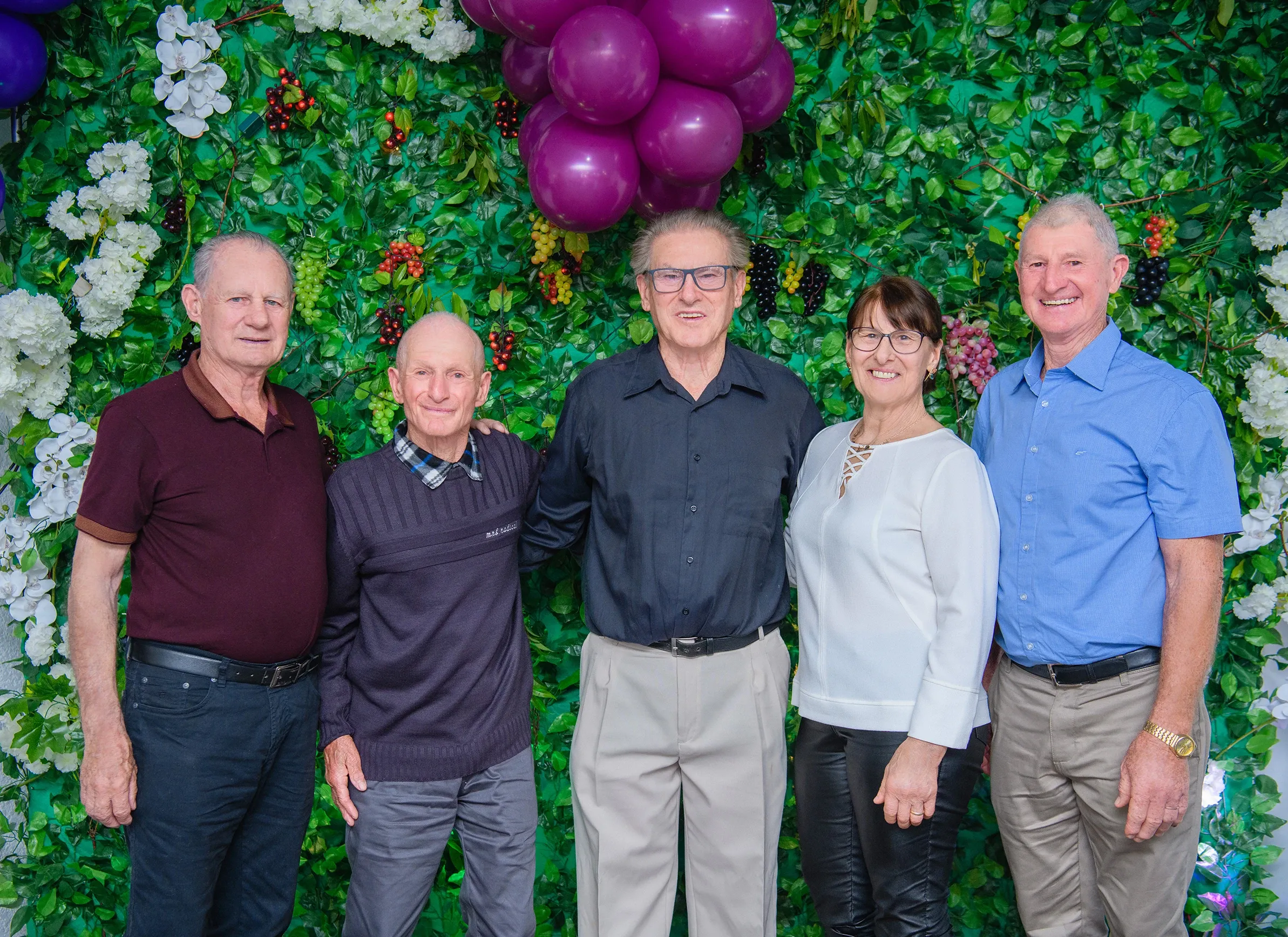 Cinco indivíduos de meia-idade e idosos, quatro homens e uma mulher, sorriem enquanto posam para uma fotografia em frente a uma parede coberta por folhagem verde e cachos de flores e bagas.