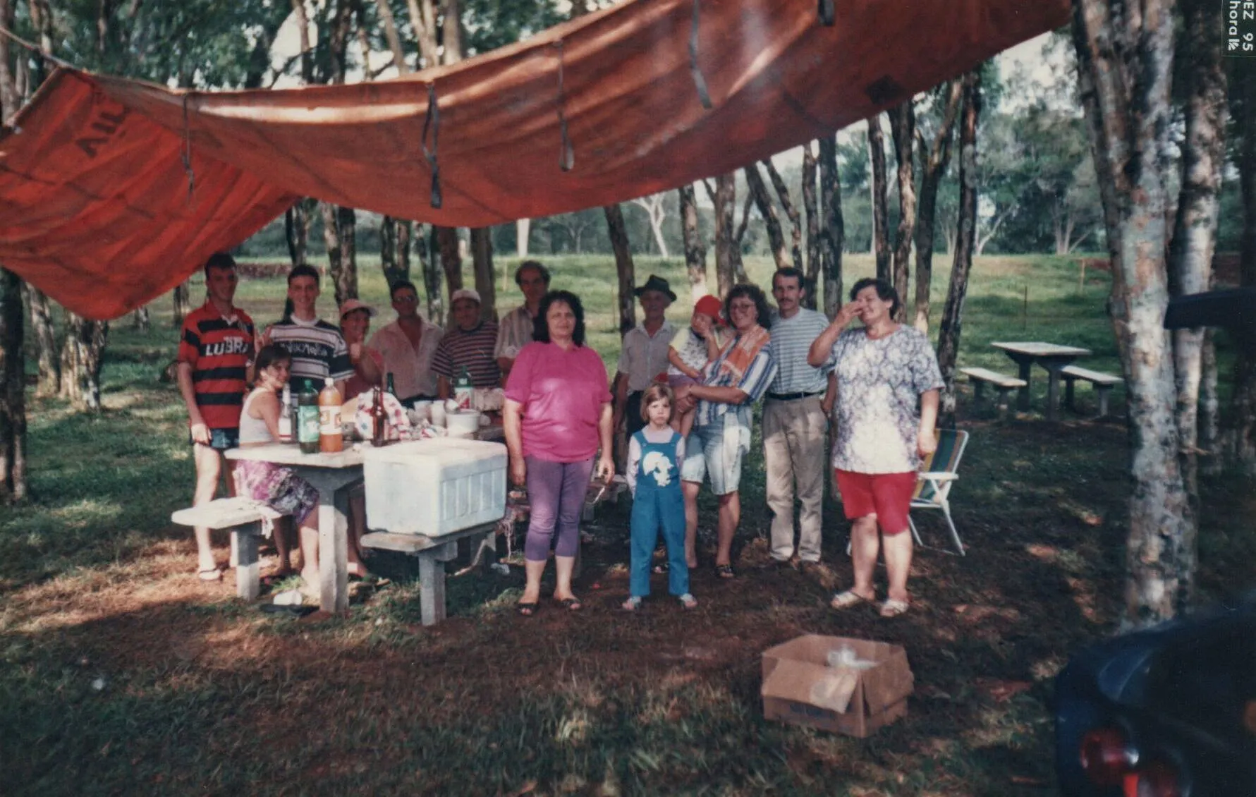 Um grupo de pessoas está reunido sob uma lona laranja estendida entre árvores, com uma mesa de piquenique cheia de comida e um cooler, desfrutando de um dia ao ar livre.
