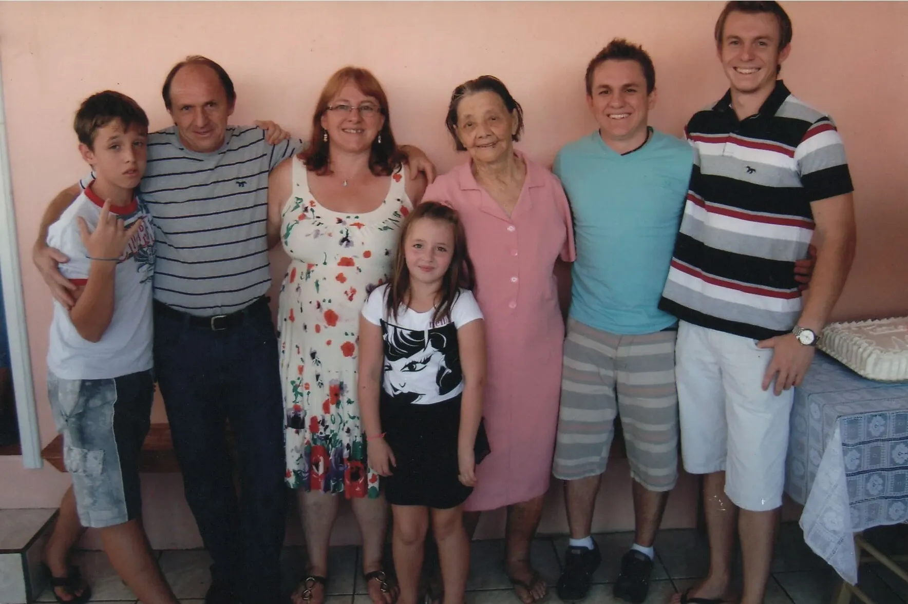 Um grupo de sete pessoas sorridentes — quatro homens, duas mulheres e uma menina — posando lado a lado em um espaço interno com uma parede rosa.