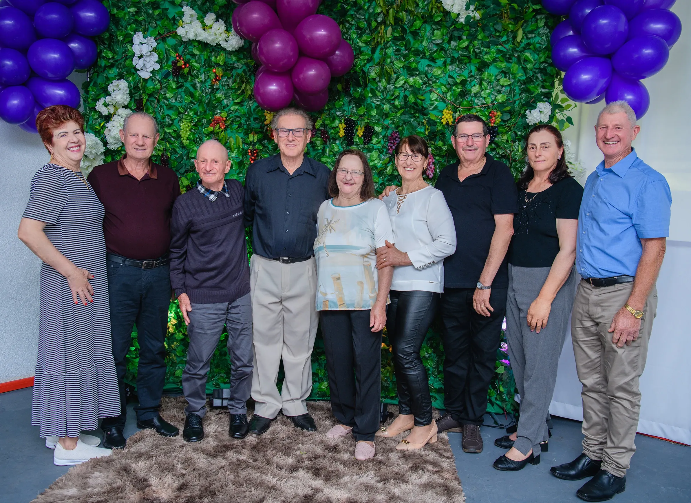 Dez adultos, cinco homens e cinco mulheres, estão alinhados e sorrindo para a câmera em frente a uma parede verde decorada com folhas, flores brancas e roxas e cachos de uvas, com balões roxos nos cantos superiores.