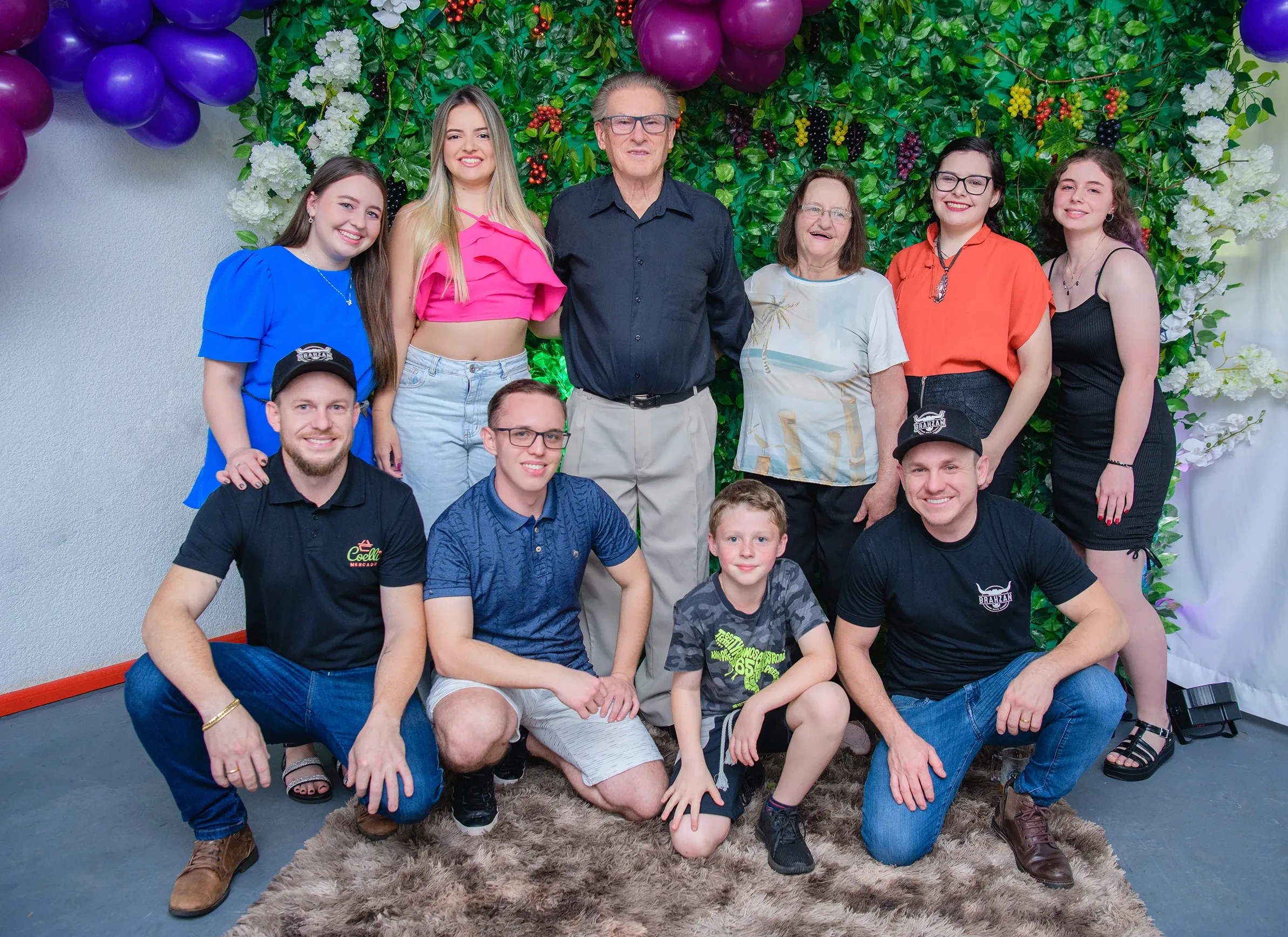 Um grupo de onze pessoas, incluindo crianças e adultos, posa para uma foto em frente a uma parede de folhagem verde adornada com balões roxos e flores brancas.