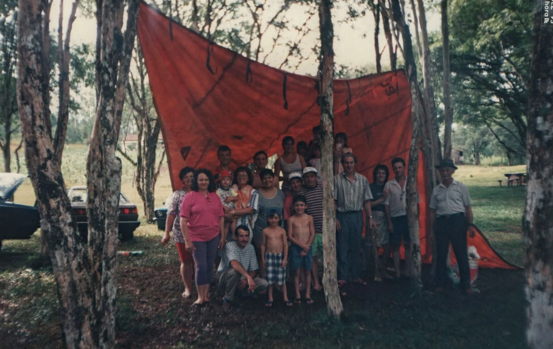 Grupo de aproximadamente 20 pessoas de várias idades está parado debaixo de uma lona vermelha esticada entre árvores, com carros estacionados e mais árvores ao fundo.