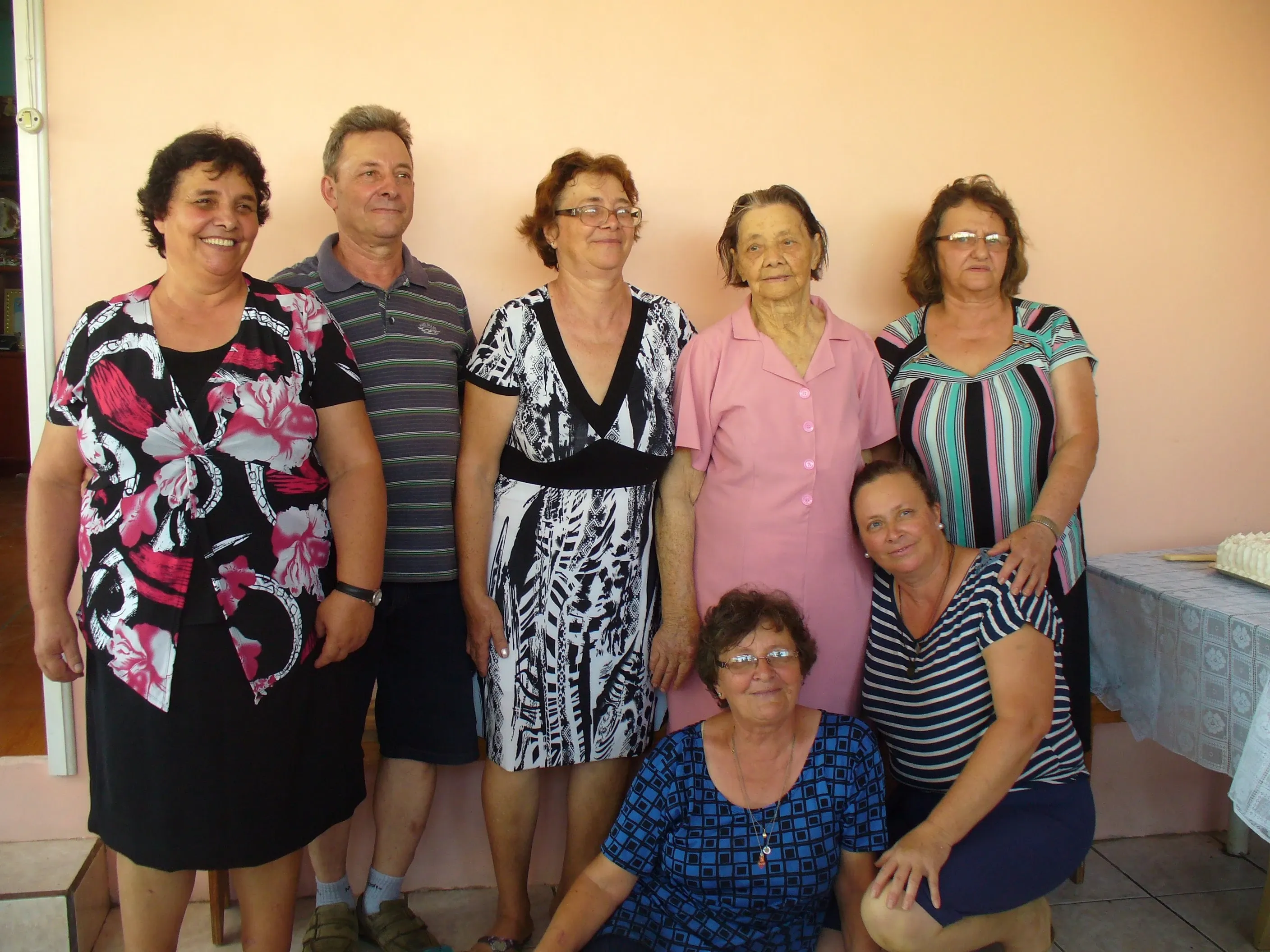 Um grupo de sete pessoas, predominantemente mulheres de meia-idade e idosas, posa para uma foto em frente a uma parede lisa de cor pêssego, com um bolo sobre a mesa no lado direito da imagem.