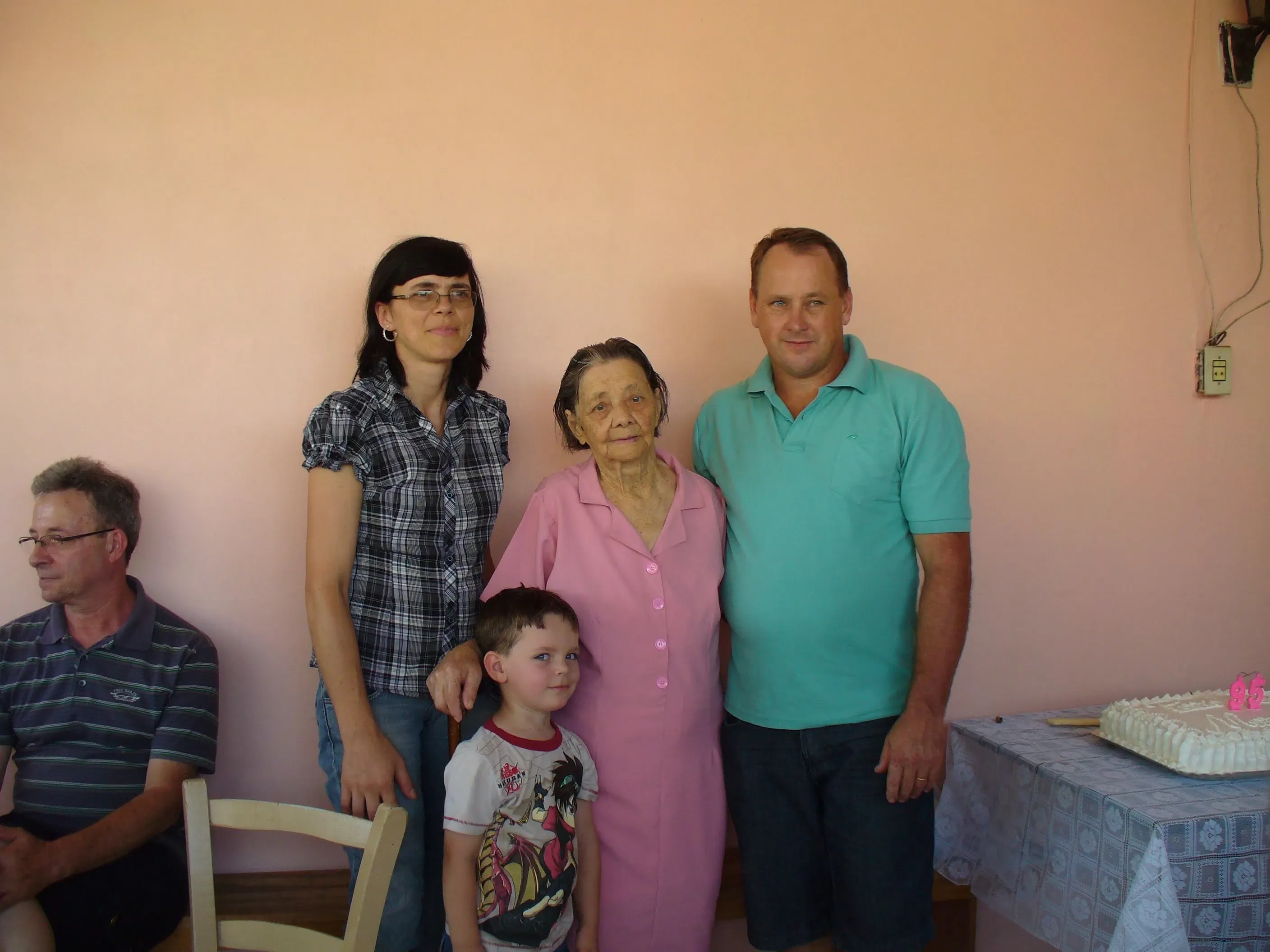 Um grupo de quatro pessoas, incluindo uma mulher mais velha em um vestido rosa e um homem em uma camisa polo azul, posa para uma foto em um interior com paredes rosa.