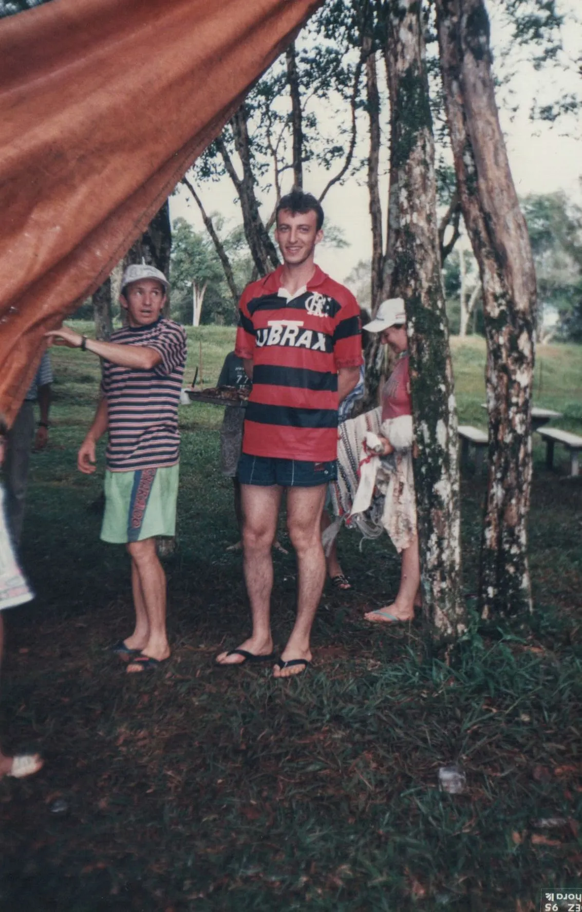 Homem em camisa listrada vermelha e preta, shorts jeans, e chinelos, parado sob uma tenda laranja ao ar livre, com árvores e outras pessoas ao fundo.