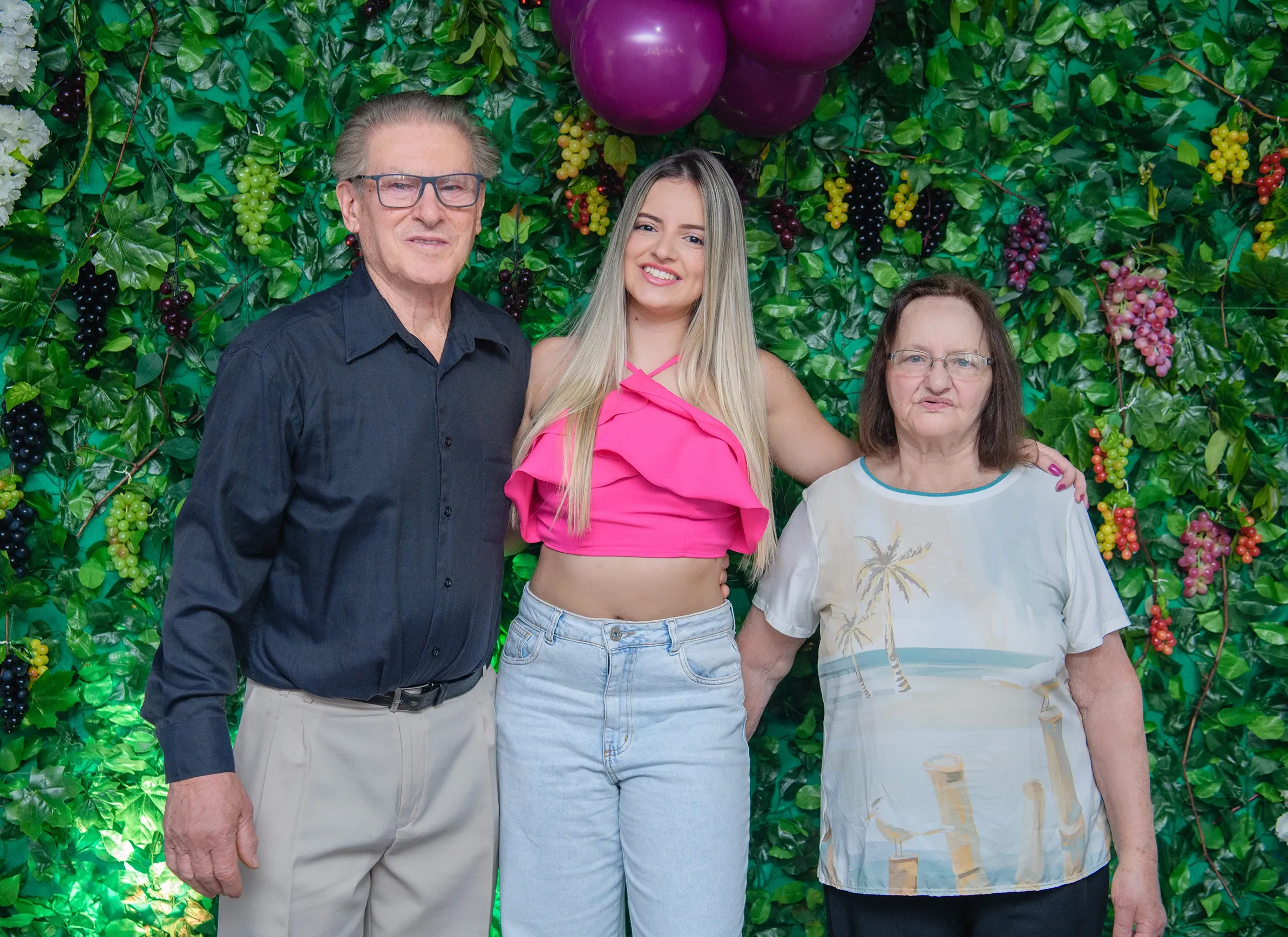 Três pessoas, um homem e duas mulheres, em pé e sorrindo contra uma parede coberta de folhas verdes e uvas, com balões roxos no canto superior direito.
