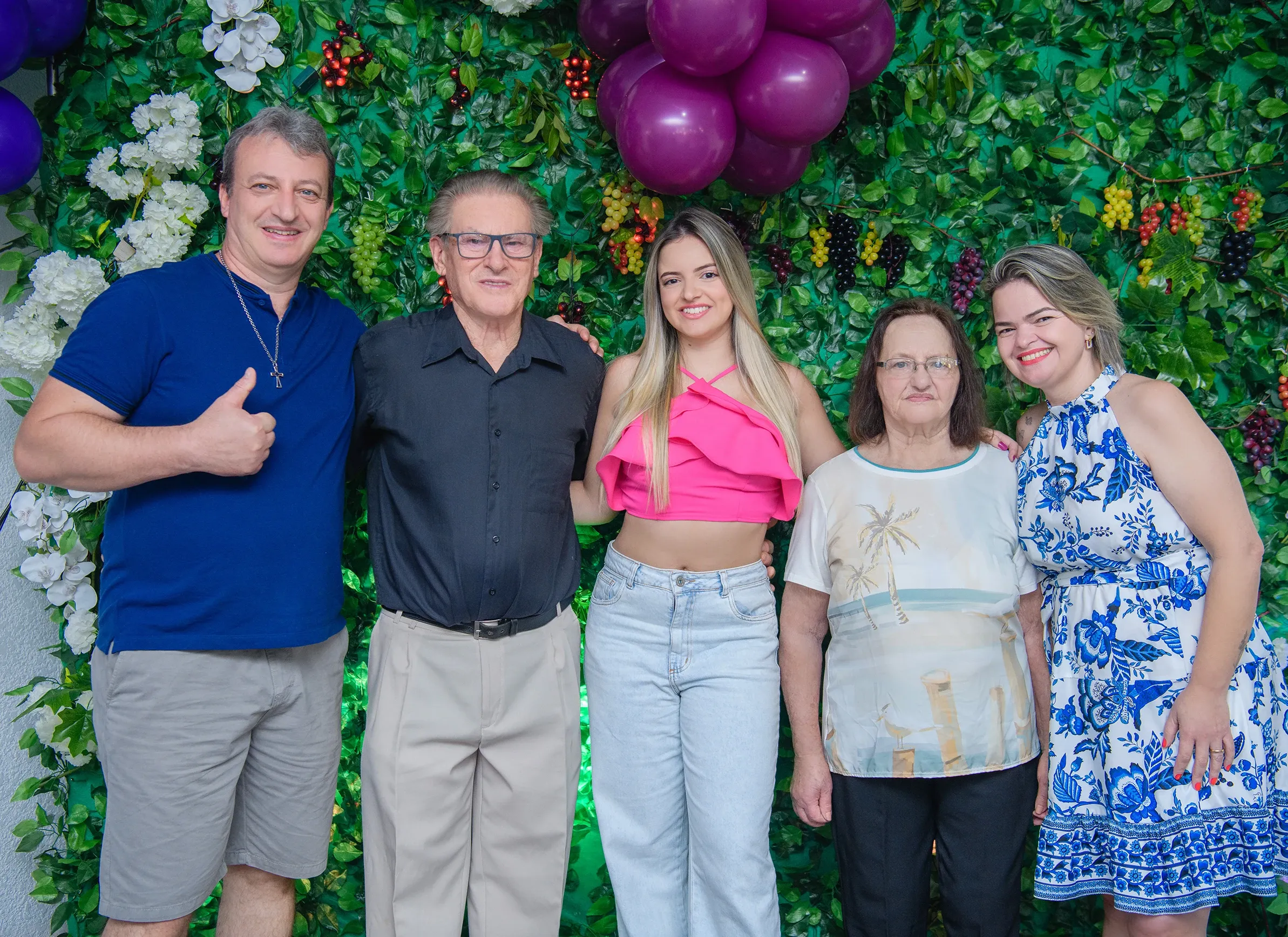 Cinco pessoas (três mulheres e dois homens) posam sorrindo na frente de uma parede coberta por folhagens verdes e frutas, com balões roxos no topo.