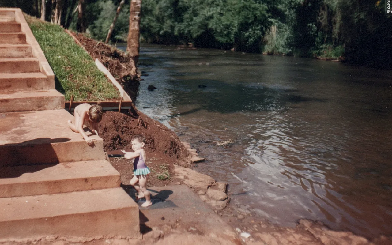 Duas crianças, uma em pé em um maiô roxo e a outra rastejando em um maiô cor da pele, estão em uma escadaria de concreto marrom na margem de um rio em uma área de floresta verde.
