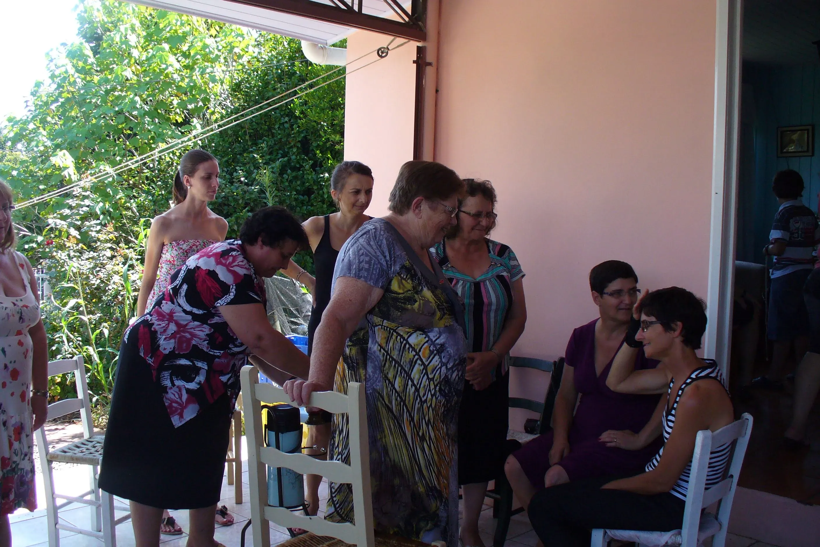 Um grupo de mulheres está reunido em um pátio externo, algumas em pé e outras sentadas em cadeiras, conversando em um dia ensolarado.
