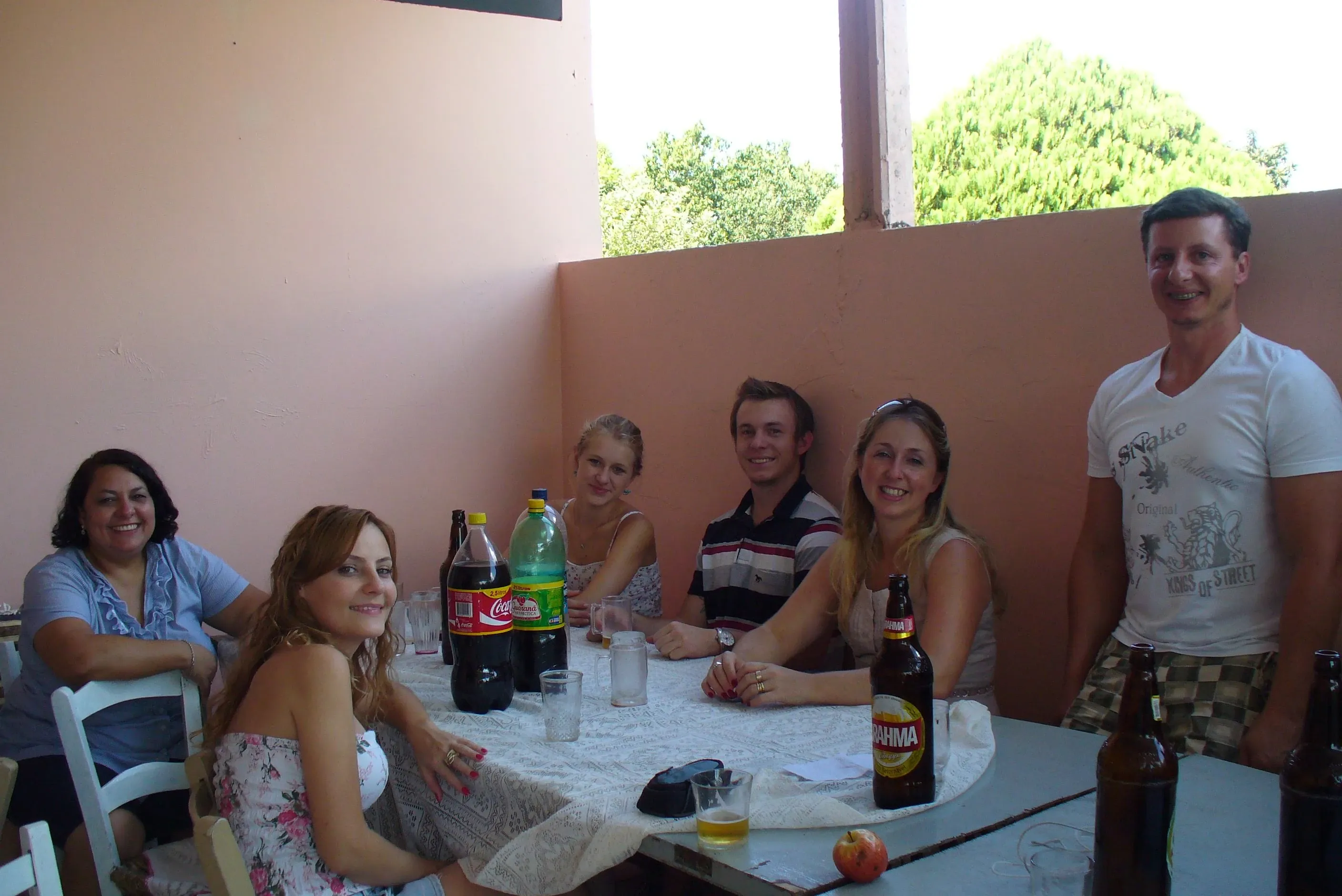 Seis pessoas sentadas ou em pé ao redor de uma mesa de pátio ao ar livre, com bebidas e comida em um dia ensolarado.