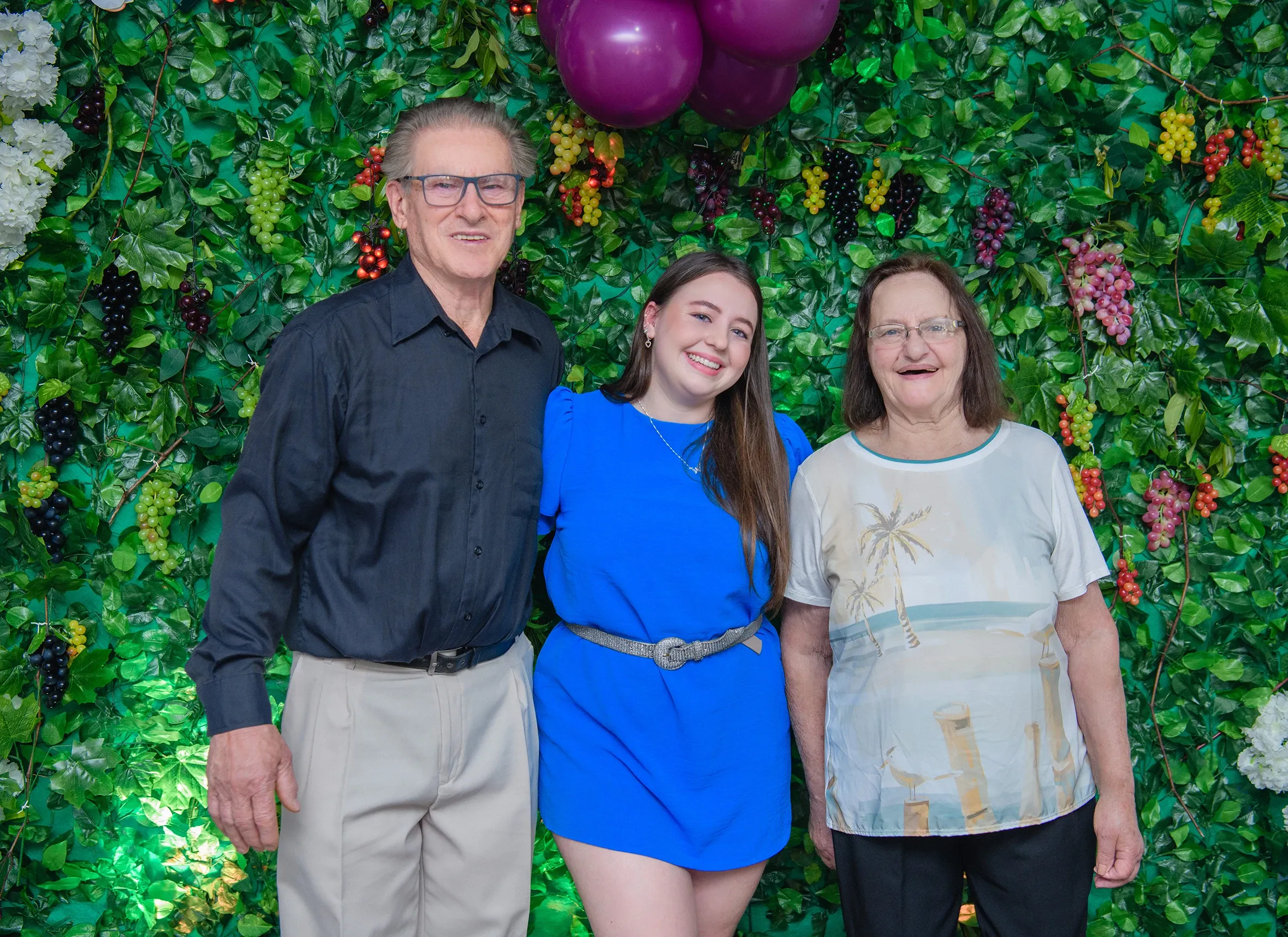 Um homem idoso, uma mulher idosa e uma jovem adulta de pé na frente de uma parede de hera, sorrindo.