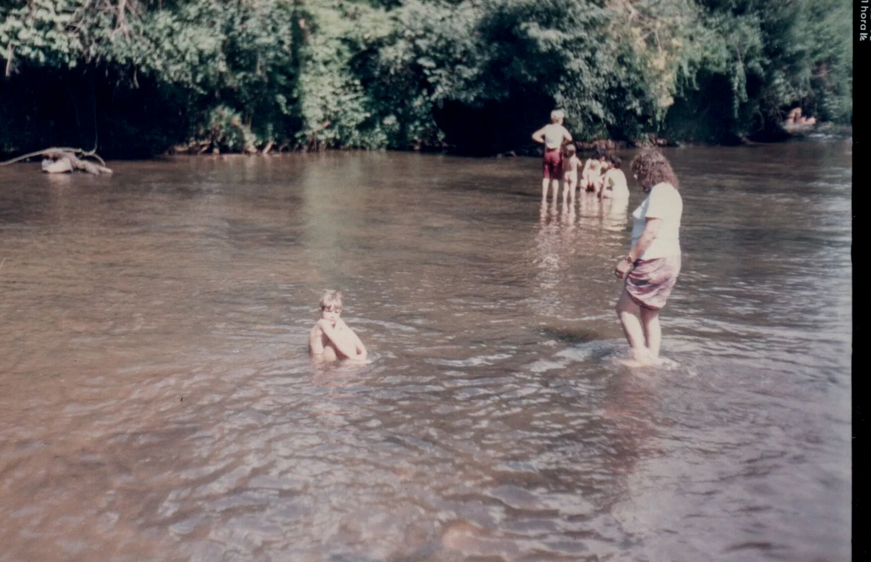Pessoas se refrescando em um rio, com uma criança sentada na água em primeiro plano e adultos em pé em segundo plano, cercados por vegetação exuberante.