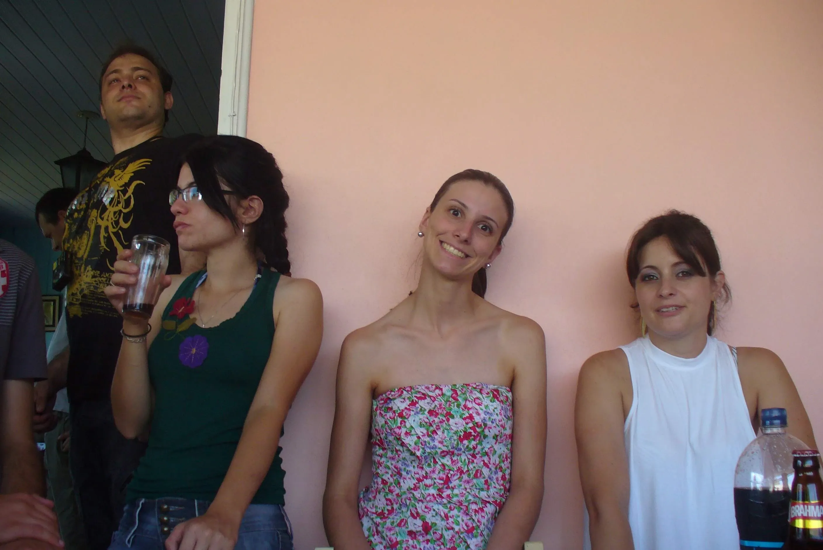 Quatro mulheres e dois homens em um ambiente fechado com paredes cor-de-rosa pêssego, um homem está no fundo olhando para cima, as três mulheres estão em primeiro plano e uma mulher está segurando um copo.