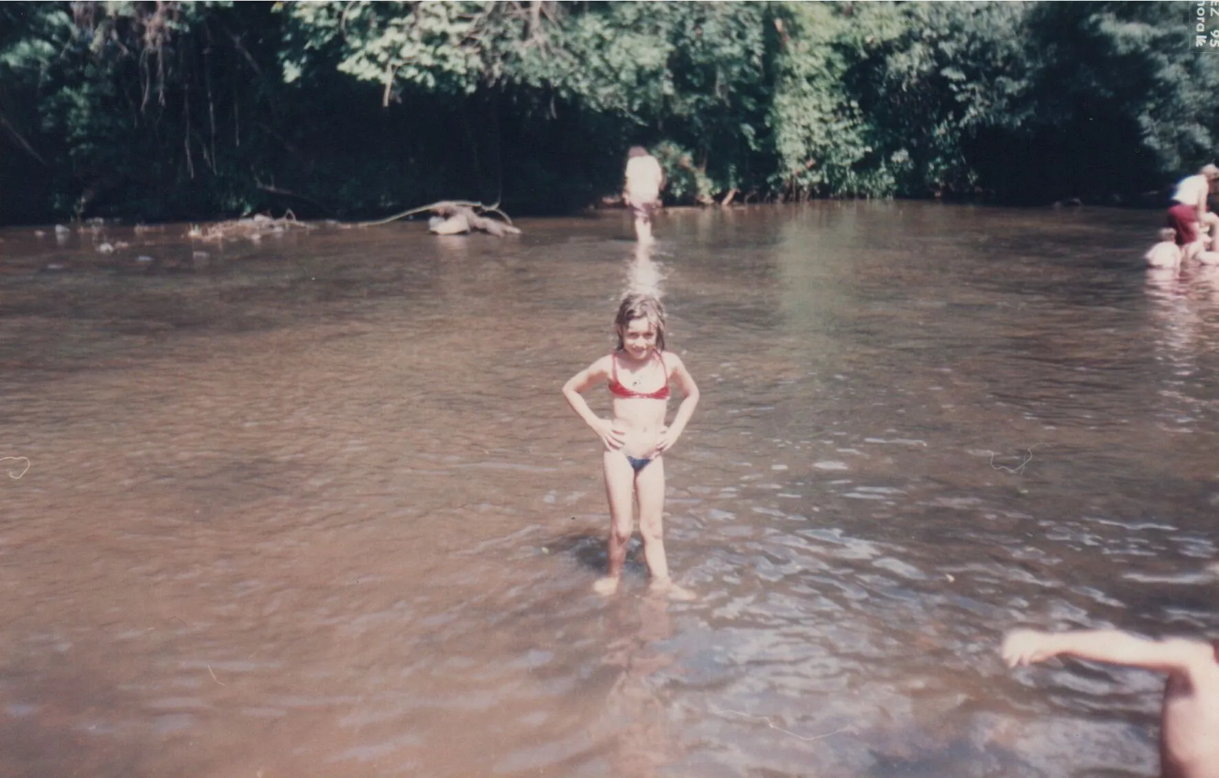 Uma garota com um biquíni vermelho e azul fica na água de um rio em um dia ensolarado, com pessoas ao fundo e uma margem arborizada.